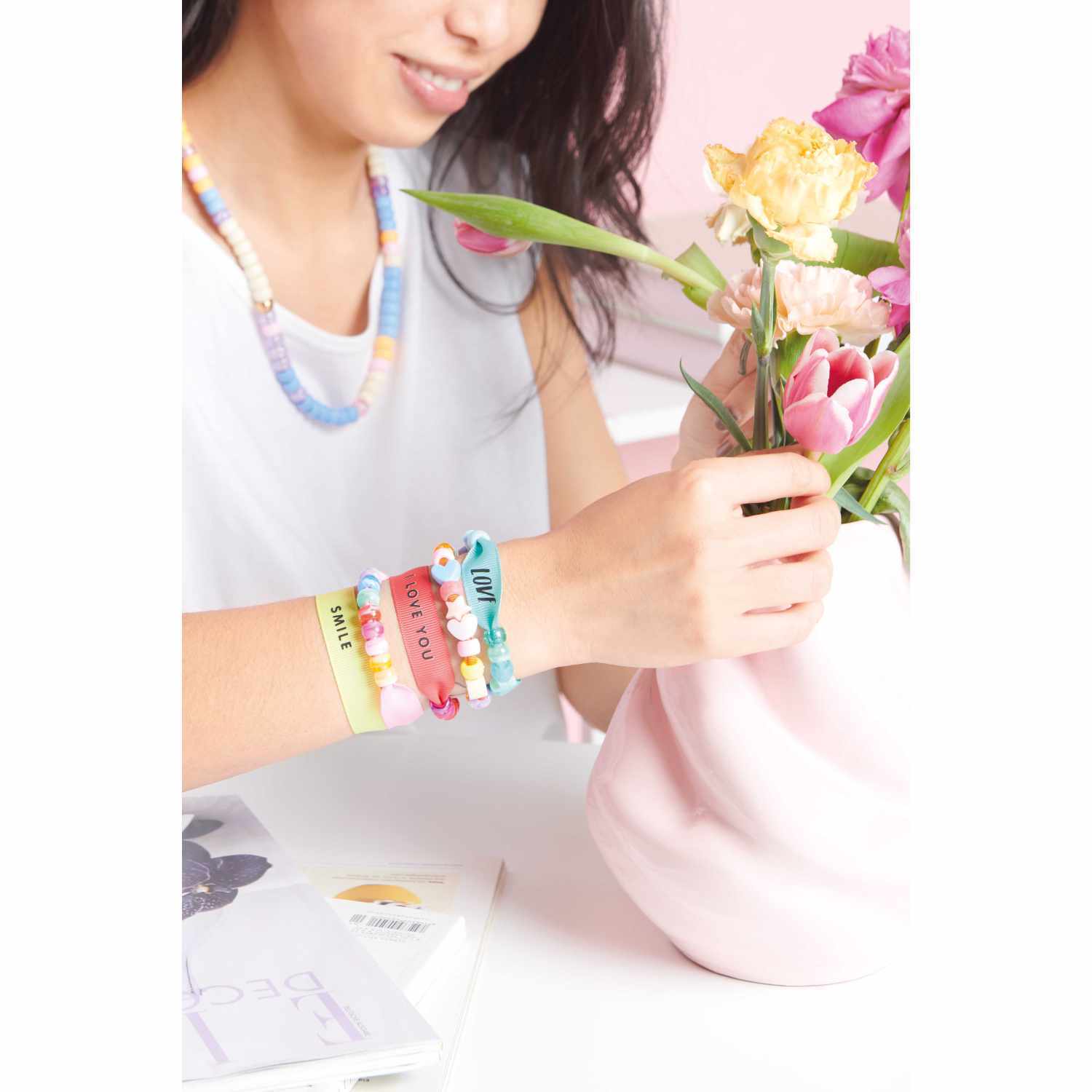 Itoshii - Ponii Beads Bügelmotiv Schriftzüge 8 Designs: Frau arrangiert Blumen in rosa Vase, trägt bunte Armbänder und Halskette.
