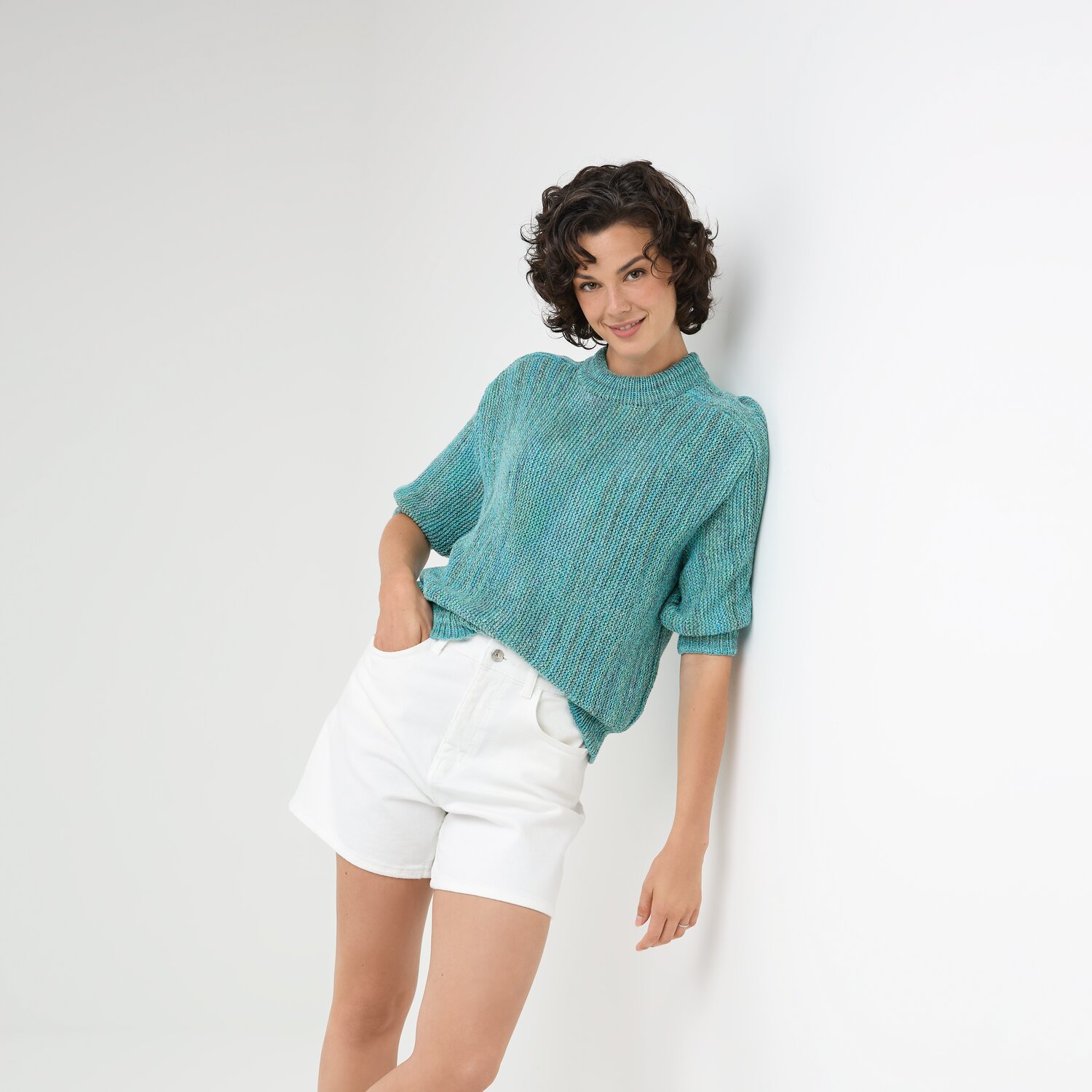 Strickset Shirt Modell 17: Frau trägt türkisfarbenes Strickshirt, lehnt an weißer Wand, kombiniert mit weißen Shorts.