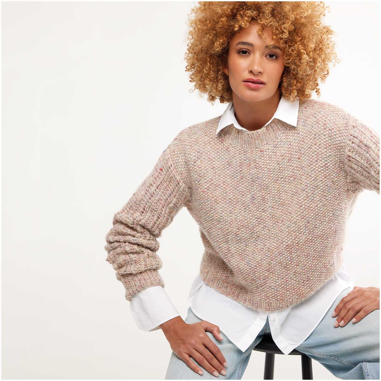 Strickset Pullover Modell 11: Frau trägt beigen Strickpullover über weißem Hemd, sitzt auf Hocker.