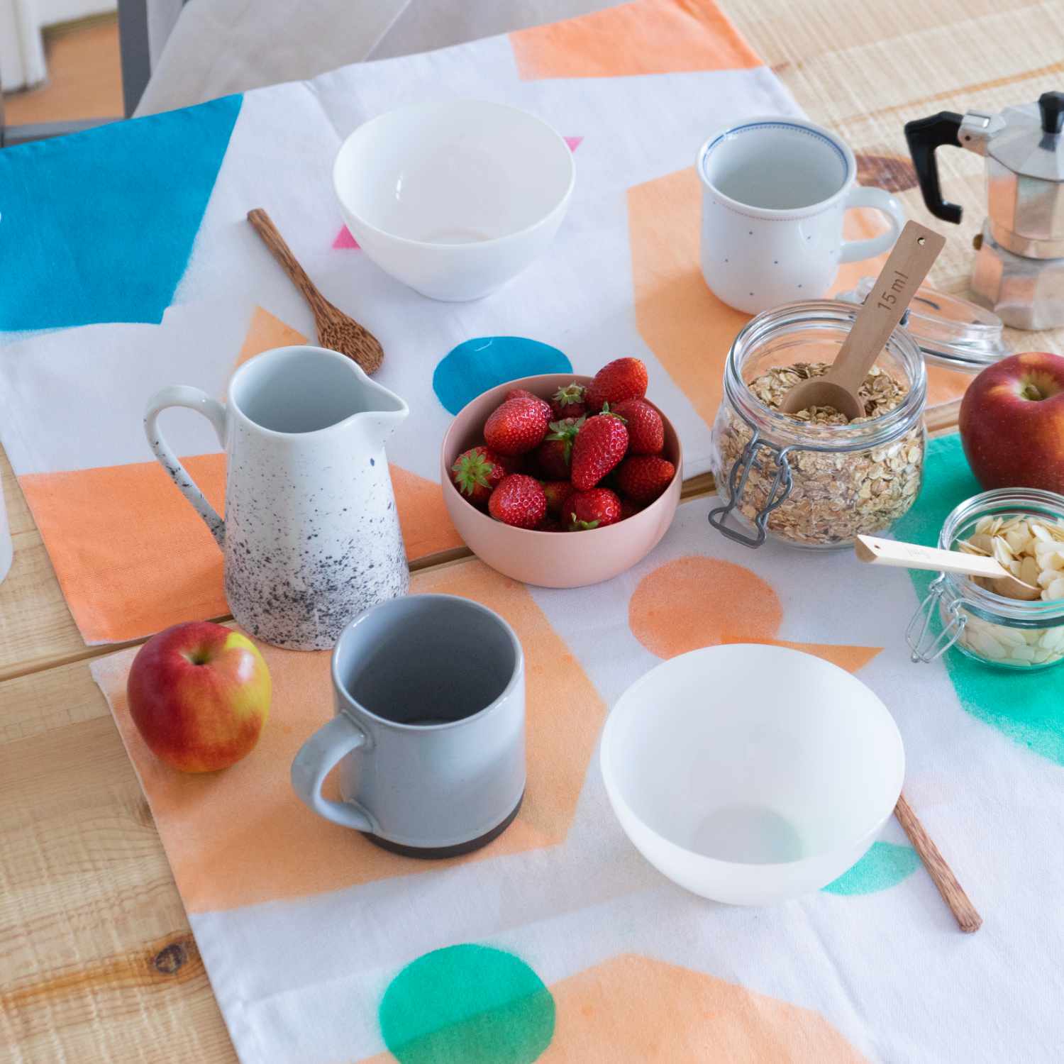 Frühstückstisch mit buntem Geschirrtuch, gefüllt mit Schalen, Tassen, Erdbeeren und Müsli im Glas.