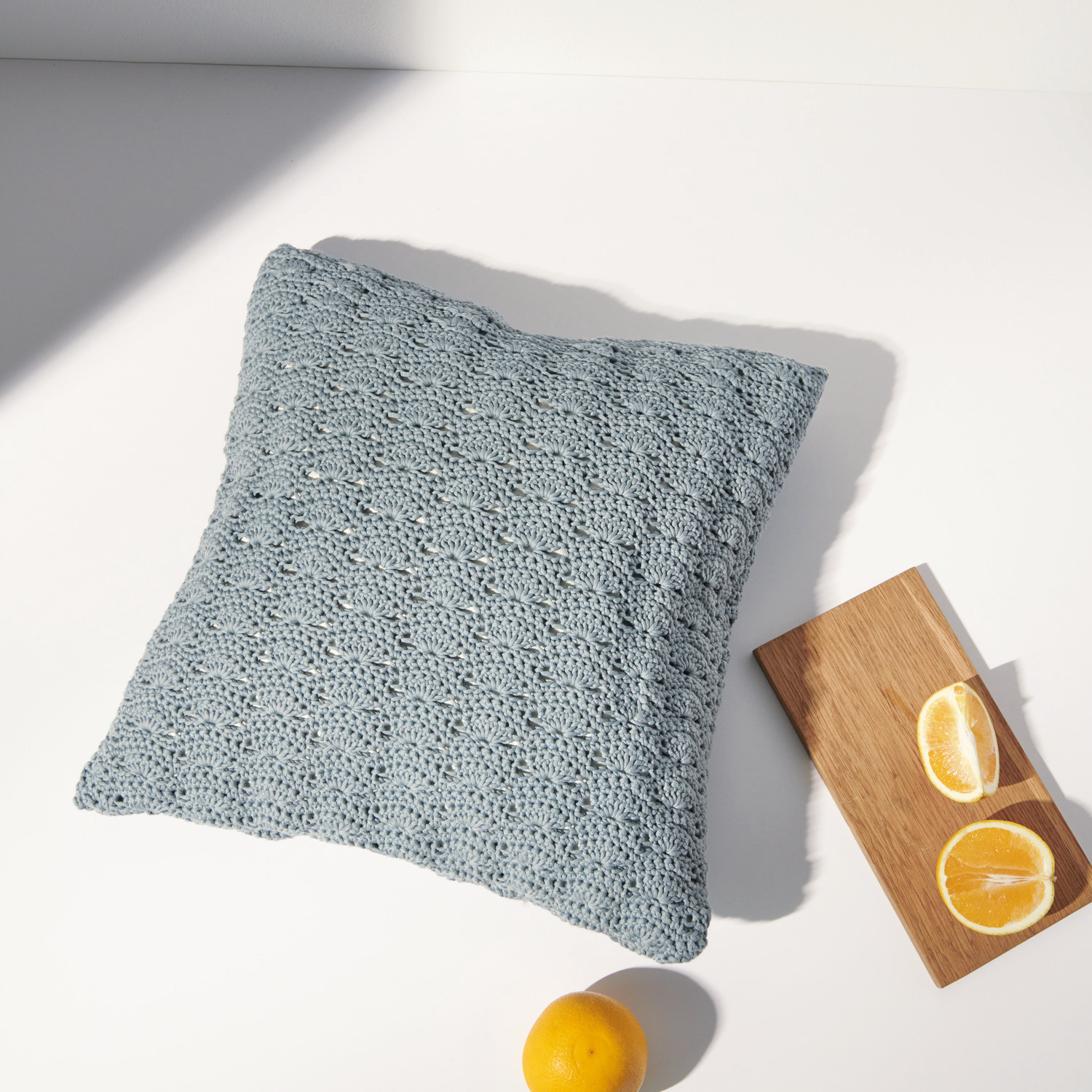 Häkelset Kissen Modell 08 aus Boho Crochet: Graues Kissen auf weißem Hintergrund, daneben Zitronenscheiben auf Holzbrett.