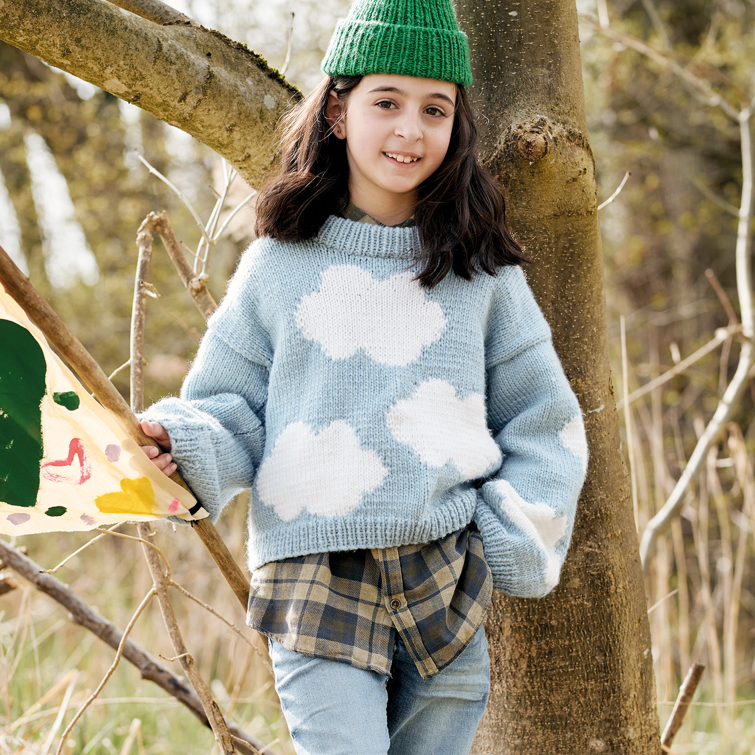 Strickanleitung Pullover aus Essentials Super aran: Mädchen im blauen Pullover mit Wolkenmotiv, grüne Mütze, im Wald mit Drachen.
