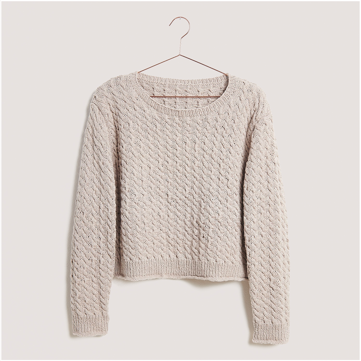 Strickset Pullover Modell 19 aus Lovewool Nr. 18: Beigefarbener Pullover mit Zopfmuster auf einem Kleiderbügel vor neutralem Hintergrund.
