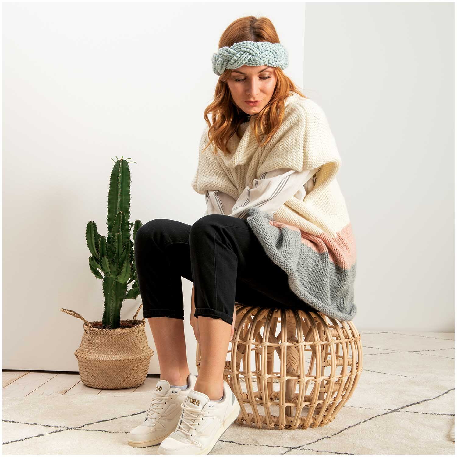Essentials Mega Wool chunky: Frau in cremefarbenem Strickponcho sitzt auf Korbhocker neben Kaktus.