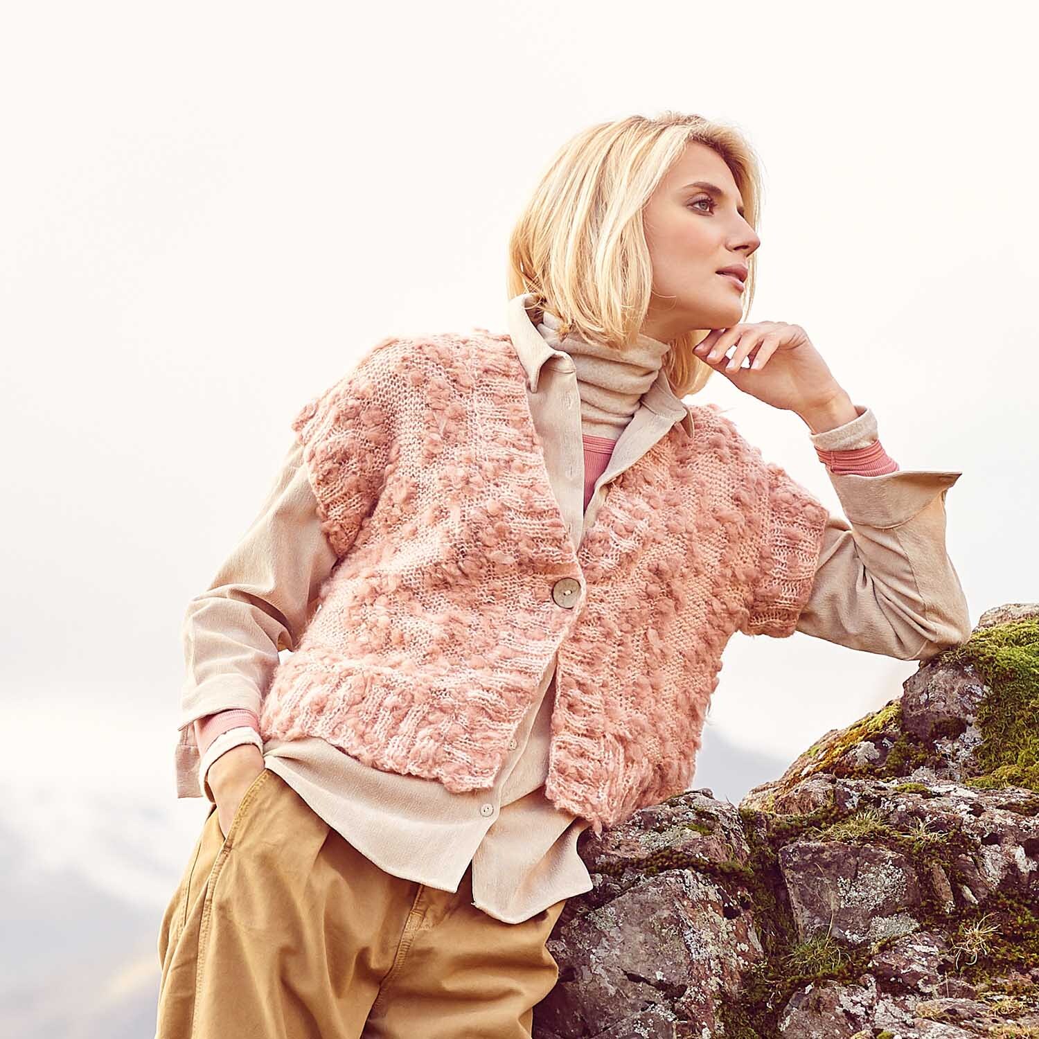 Strickanleitung Weste aus Fashion Alpaca Vibes: Frau in rosa Strickweste, beige Bluse und Hose, lehnt an Felsen in alpiner Landschaft.