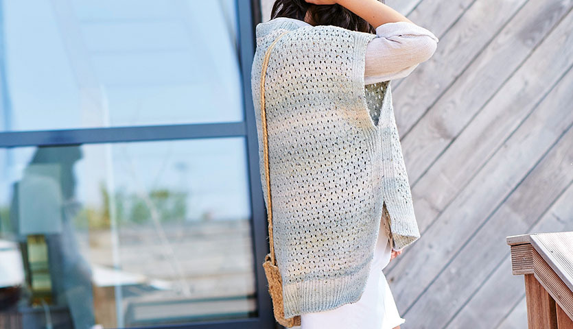 Strickanleitung Oversized Shirt aus Fashion Cotton Light & Long Tweed dk: Frau trägt gestricktes, helles Oberteil vor moderner Holzfassade.
