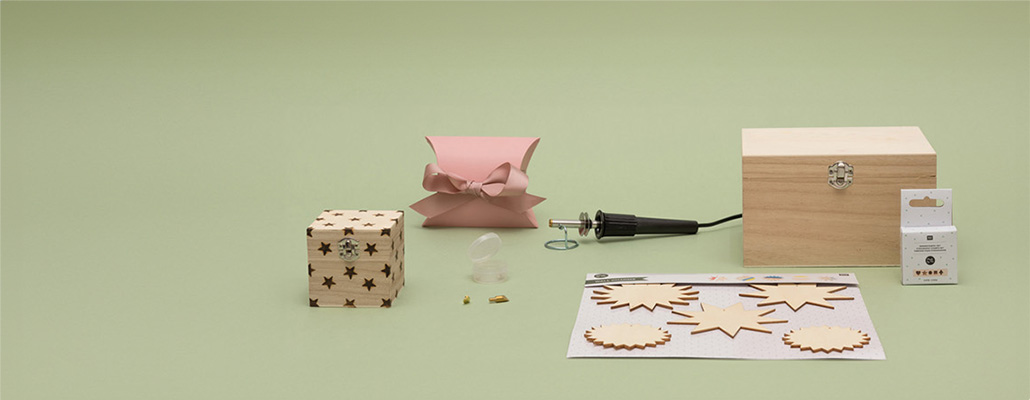 Ein Brandmalkolben mit Holzanhängern, einer großen Holzkiste und einer kleinen Holzkiste mit kleinen Sternen, sowie eine pinke Geschenkschachtel. Fotosetting grüne Wand.