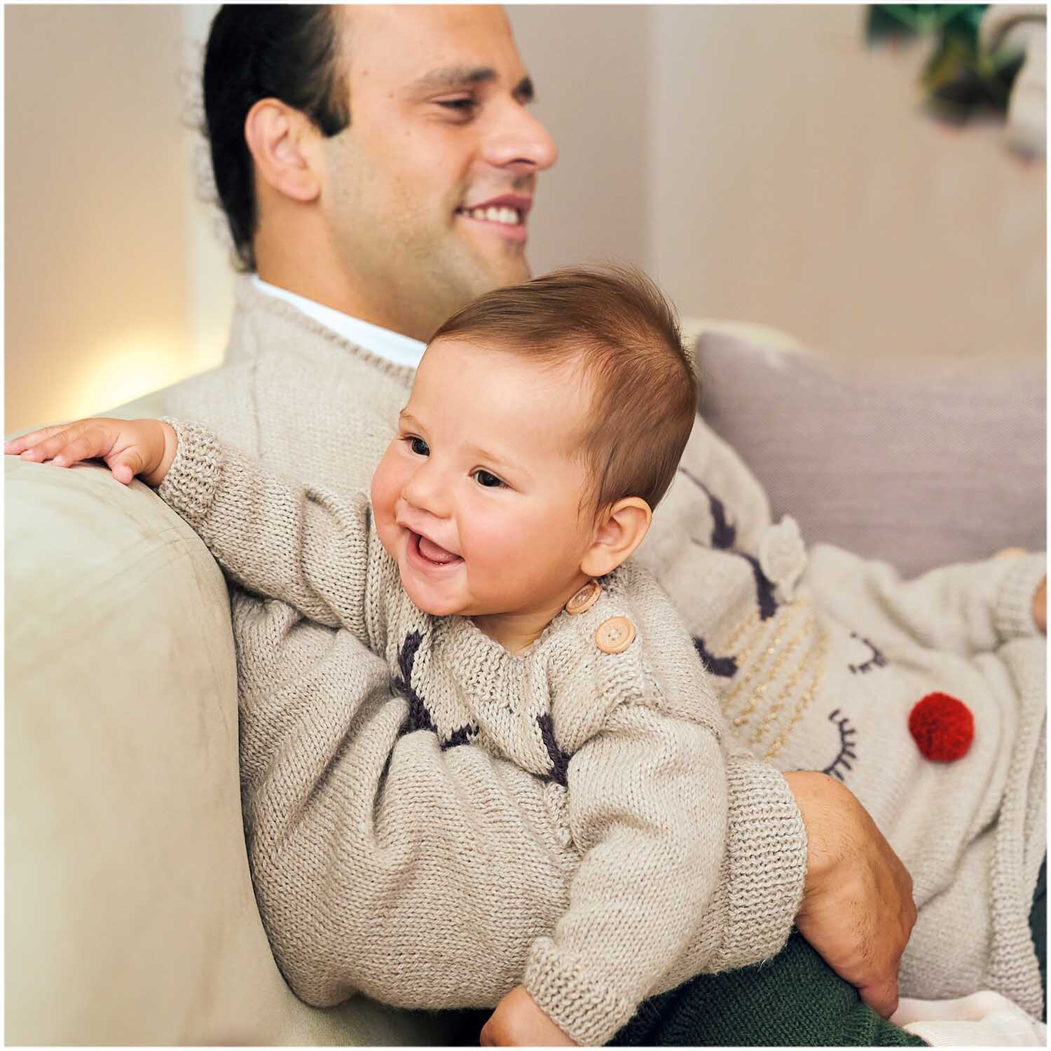 Essentials Merino Plus dk: Vater und Baby in gestrickten Pullovern aus weicher Wolle, lächelnd auf dem Sofa.