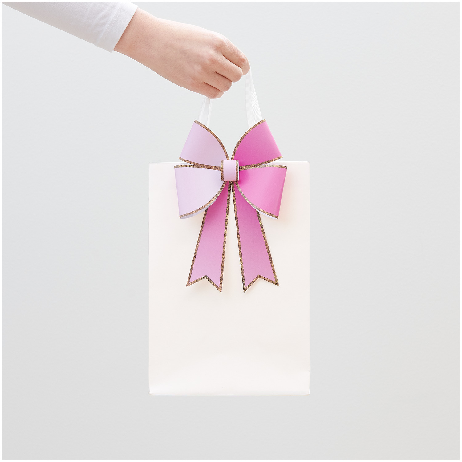 Paper Poetry Bastelpapierblock Put a Bow on It: Hand hält weiße Tasche mit rosa Schleife, 12 Blatt von Rico Design.