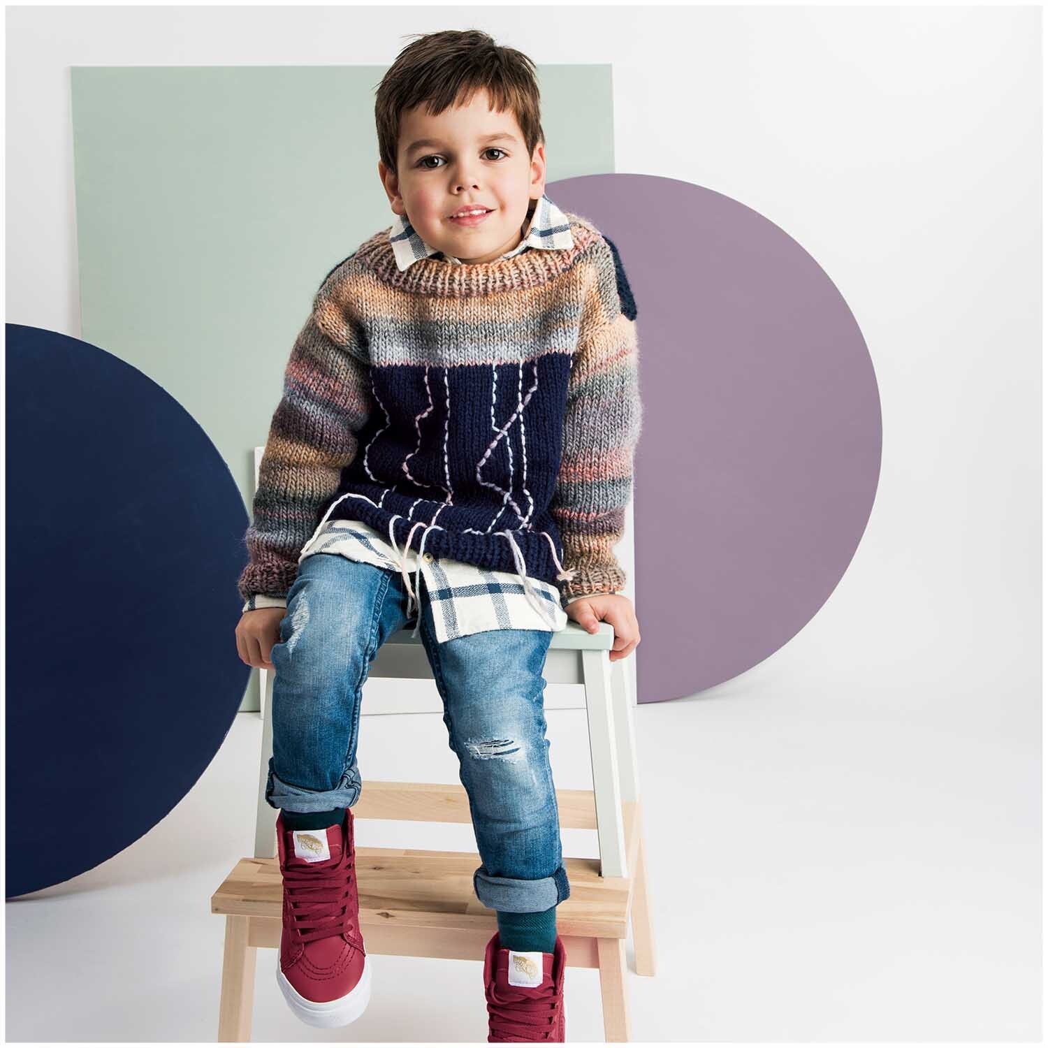 Creative Melange chunky: Ein Junge trägt einen gestrickten Pullover in Grau-Rosa von Rico Design, sitzt auf einem Stuhl vor bunten Kreisen.