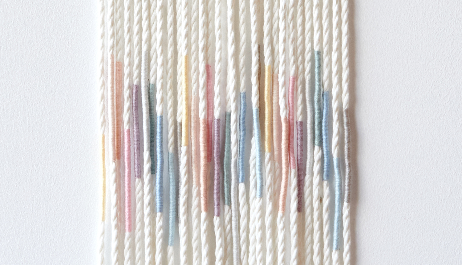 Bastelanleitung Sticktwist-Wanddeko Pastell: Wanddeko aus weißem Garn mit pastellfarbenen Akzenten.