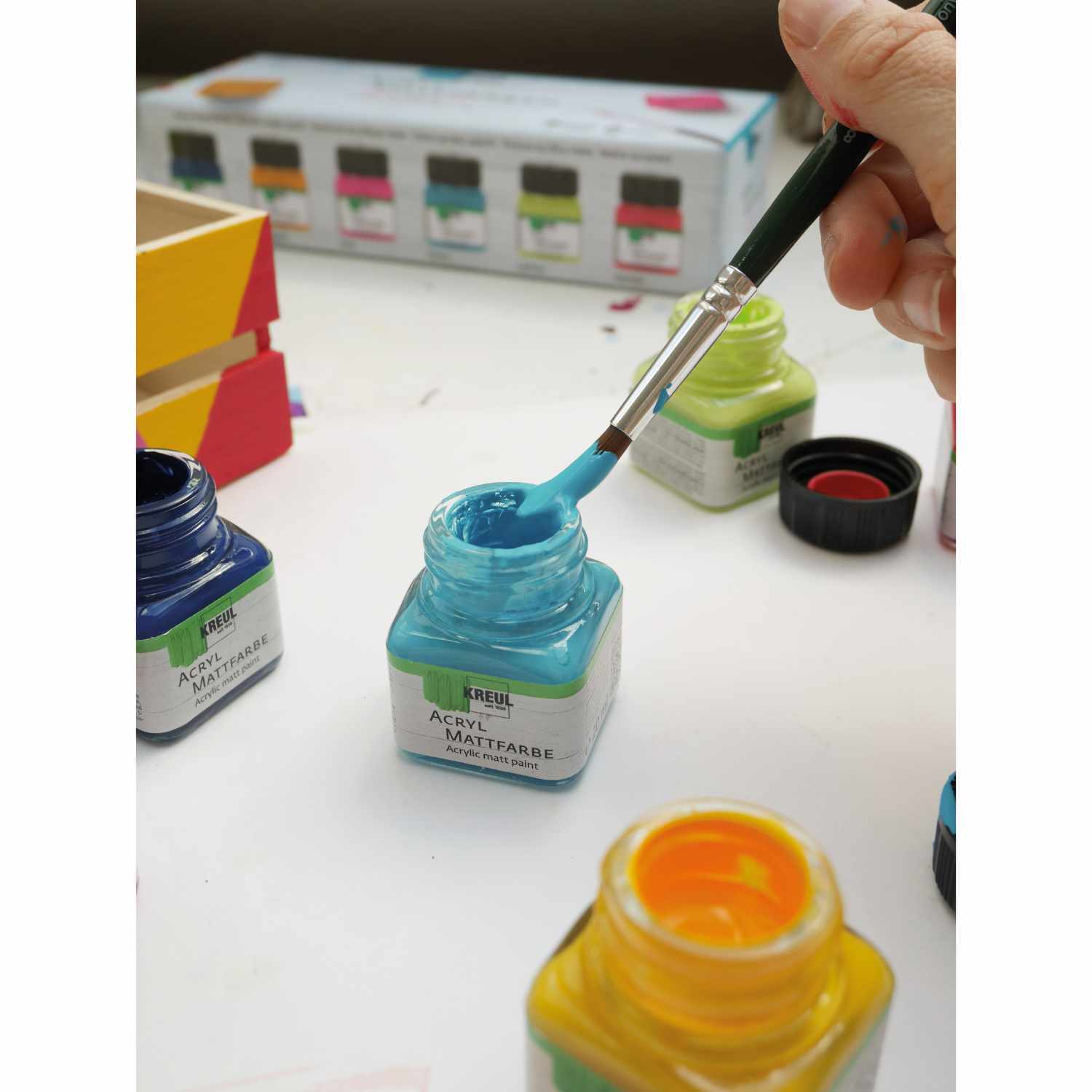 Acryl Mattfarben Set Color Living 6x20ml: Hand hält Pinsel in blauer Farbe, umgeben von bunten Farbtöpfen.