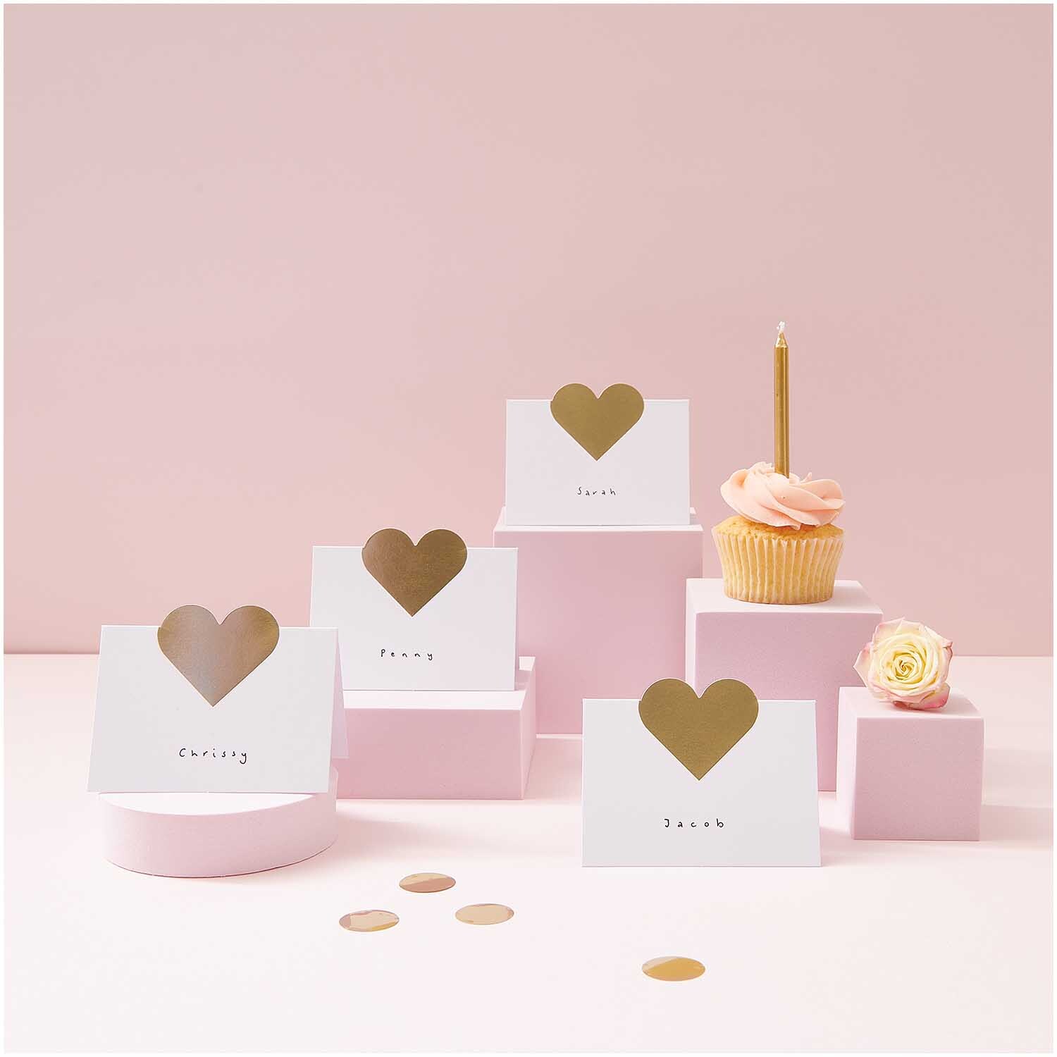 Paper Poetry Tischkarten mit Herzmotiv auf rosa Podesten, dekoriert mit Cupcake und Blume.