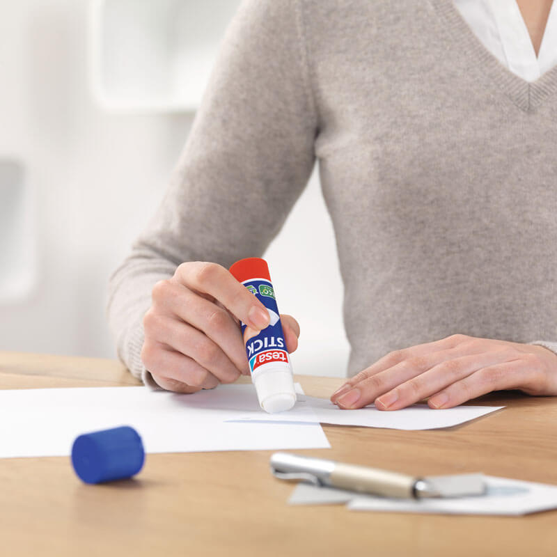 Stick Klebestift von tesa in der Hand einer Person, die Papier klebt. Tisch mit Stift und Deckel.