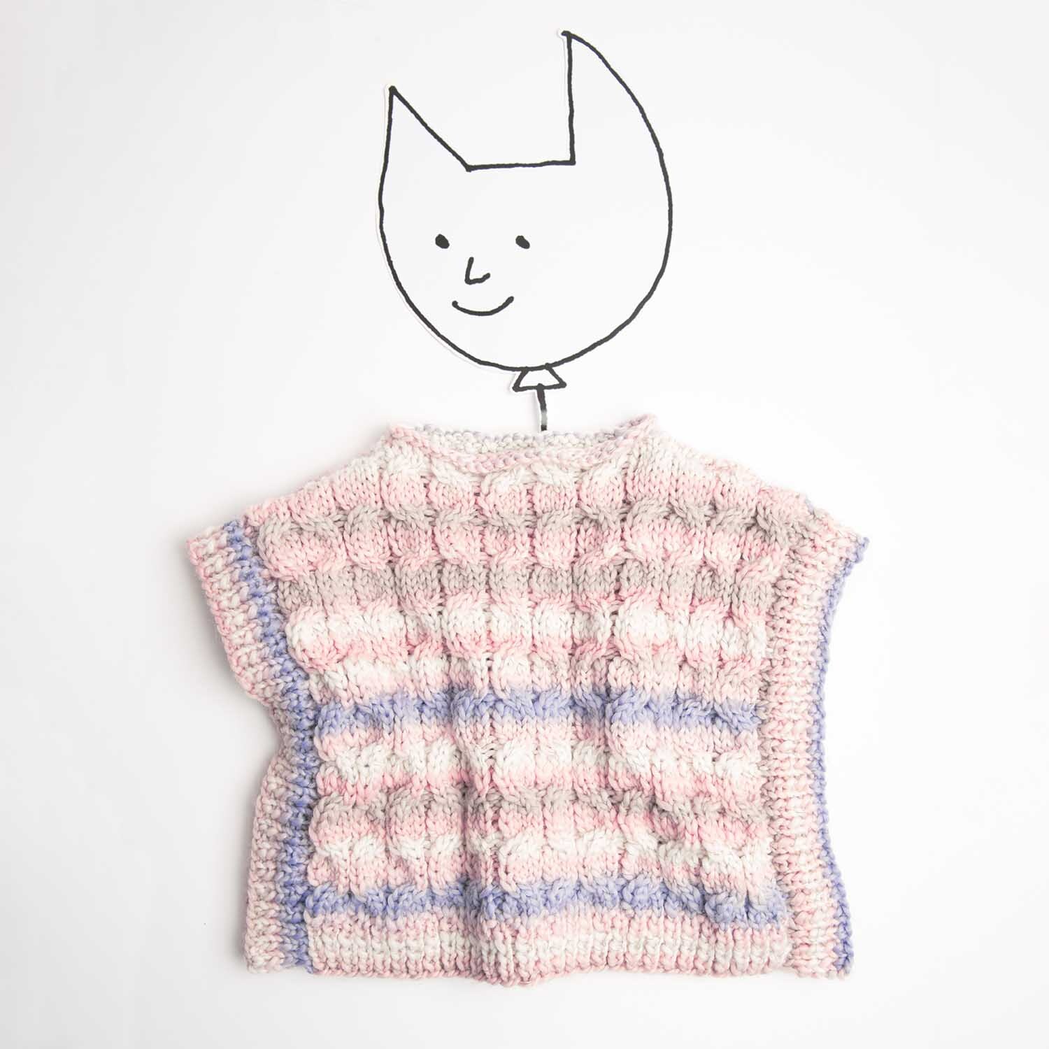 Strickanleitung Poncho aus Creative Glühwürmchen Print: Pastellfarbener Poncho mit Zopfmuster auf weißem Hintergrund, illustriert mit Katzenkopf.