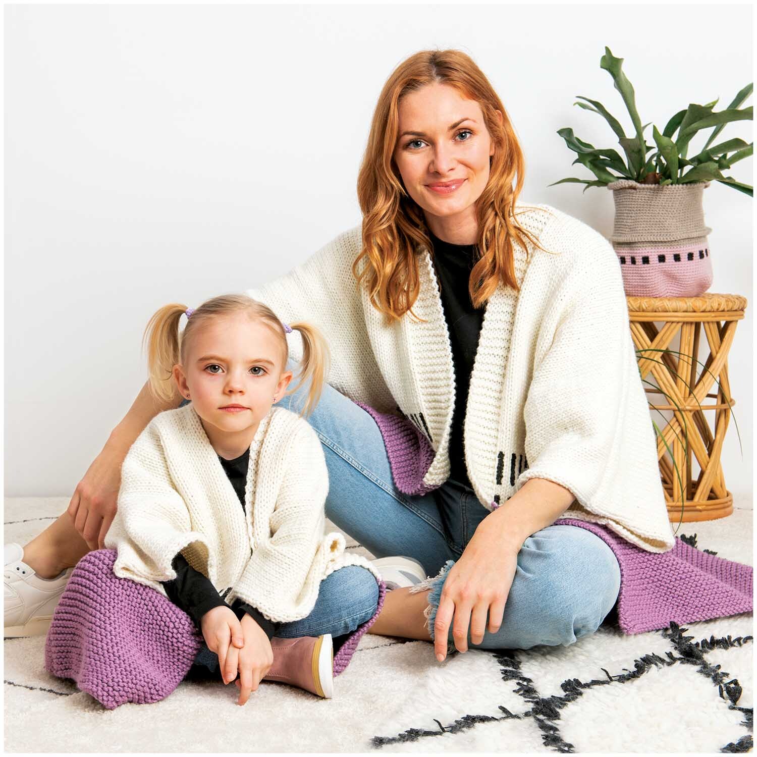 Essentials Mega Wool chunky: Mutter und Tochter in weißen Strickjacken, auf lila Decke sitzend.