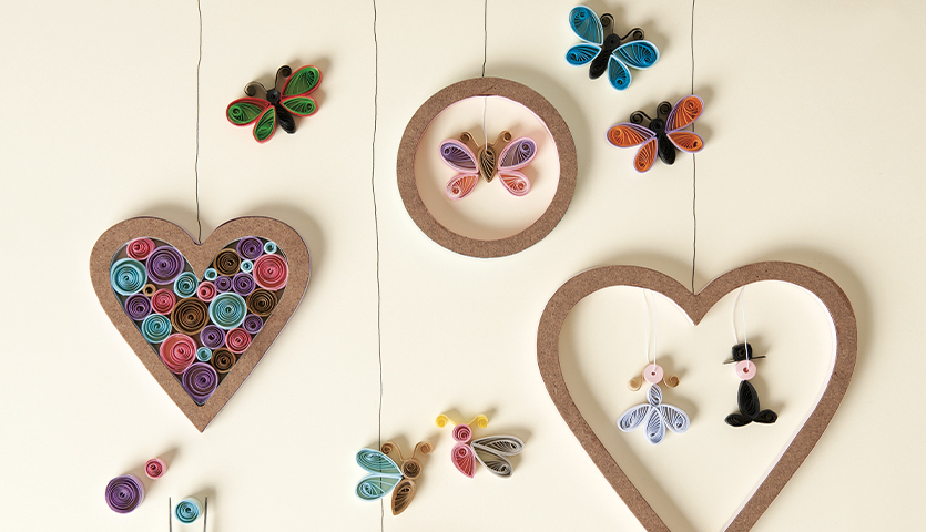 Bastelanleitung Quilling: Bunte Papierkunstwerke in Herz- und Schmetterlingsform an Fäden aufgehängt.