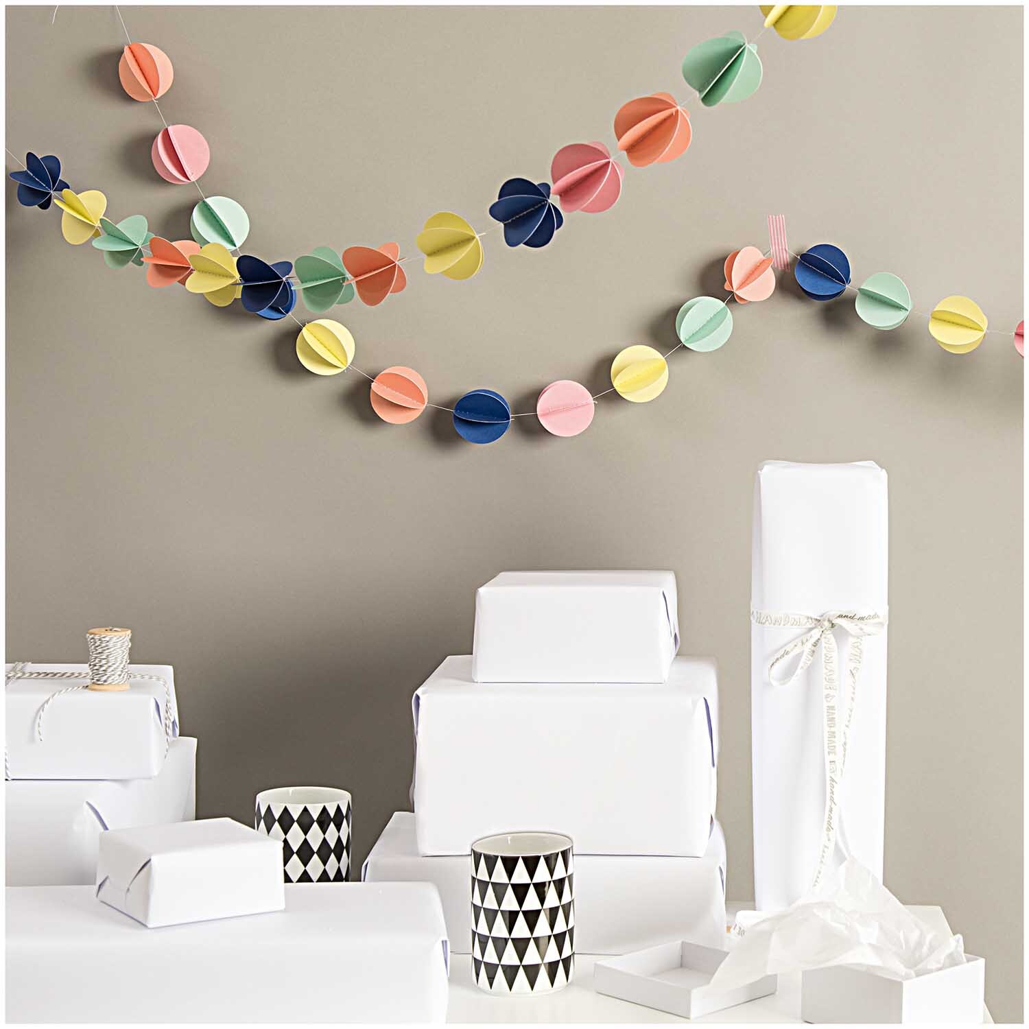Girlande Bälle mehrfarbig 1,5m von Rico Design schmückt eine Wand über weißen Geschenkboxen.