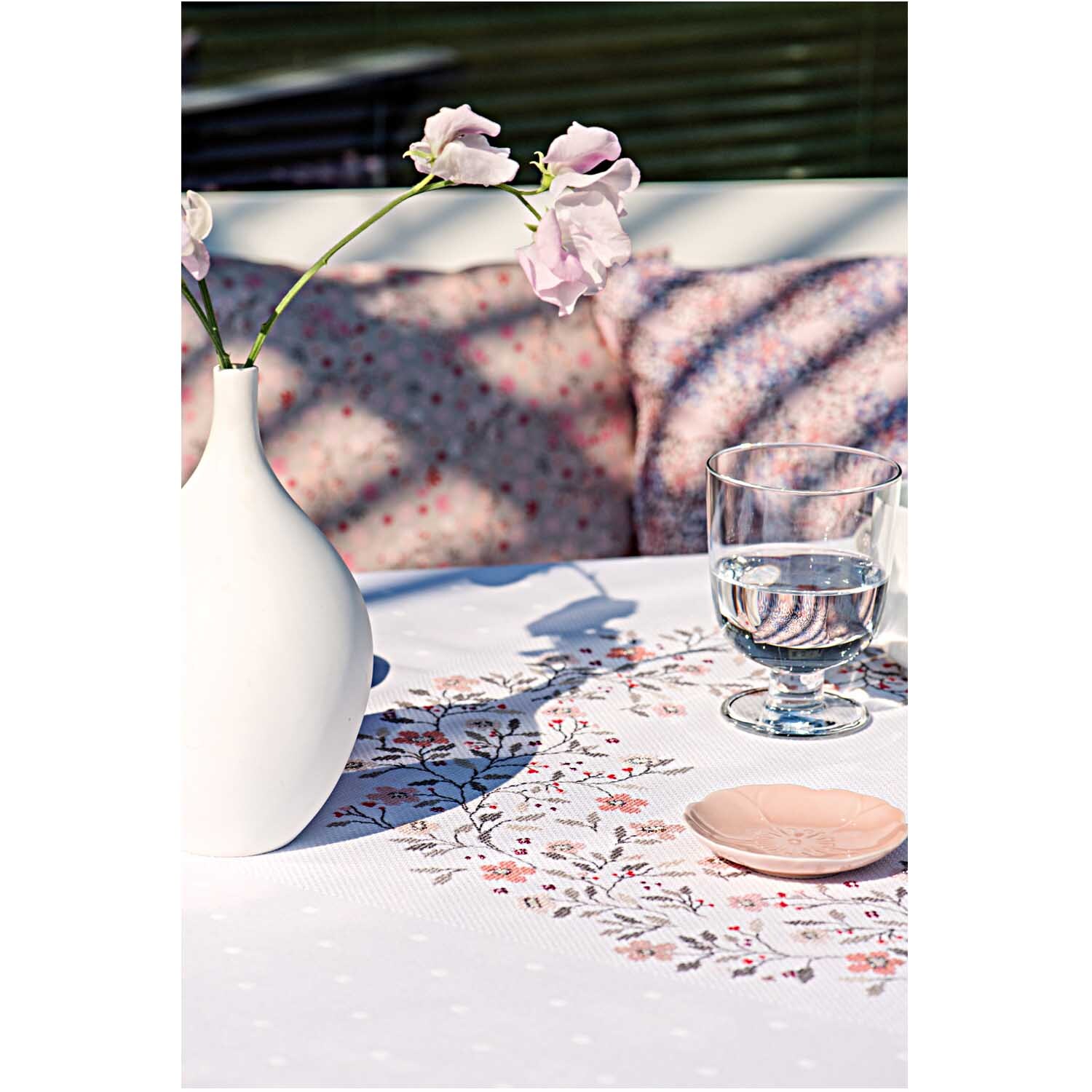 Stickpackung Tischdecke Streublümchen 90x90cm von Rico Design auf Tisch mit Vase, Glas und rosa Blüten.