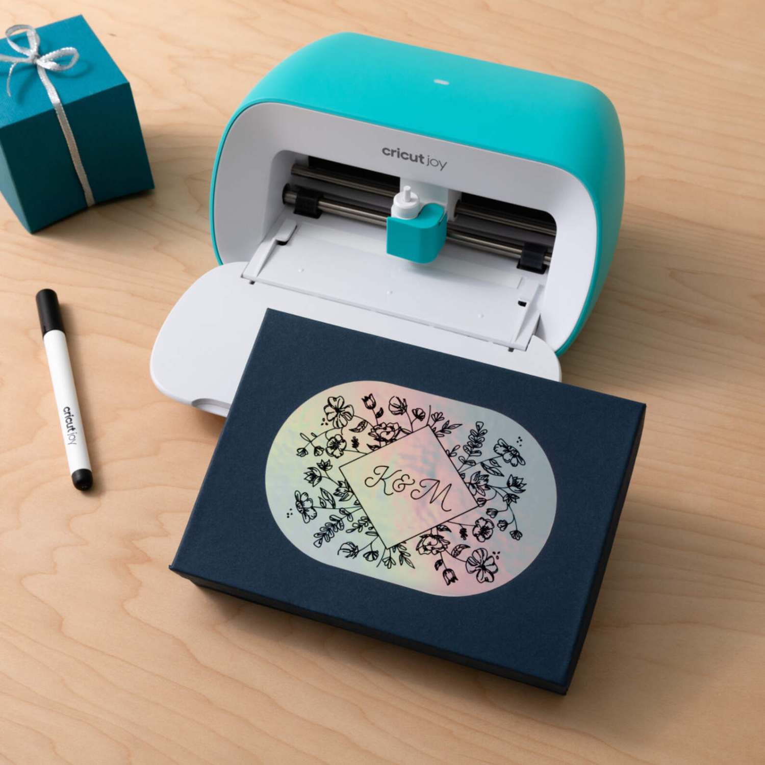Joy Smart Vinyl Permanent Writable, Cricut-Maschine erstellt personalisierte Karte auf Holzoberfläche. Daneben ein Geschenk und ein Stift.