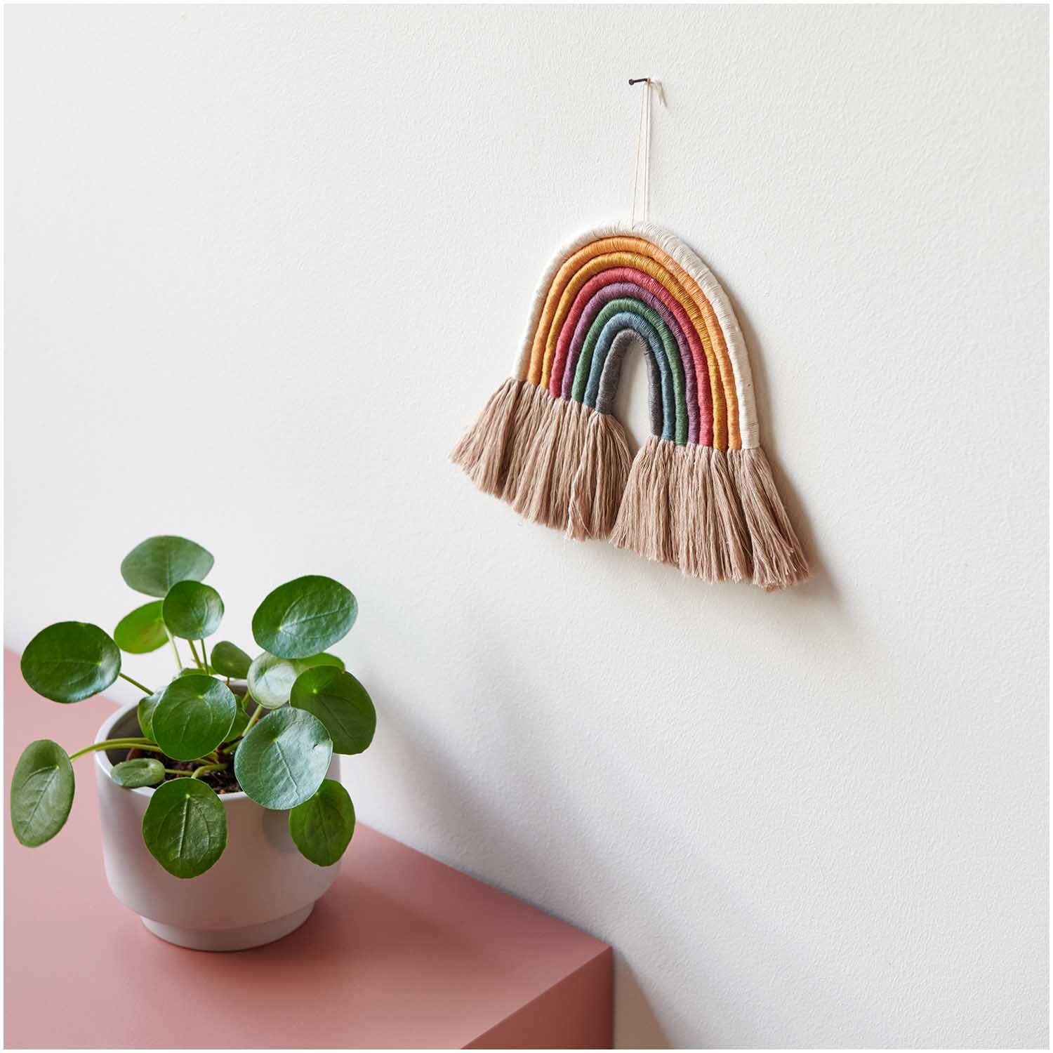 Stickgarn Set Earthy: Wanddeko mit buntem Regenbogen aus Garn, daneben grüne Pflanze auf rosa Tisch.