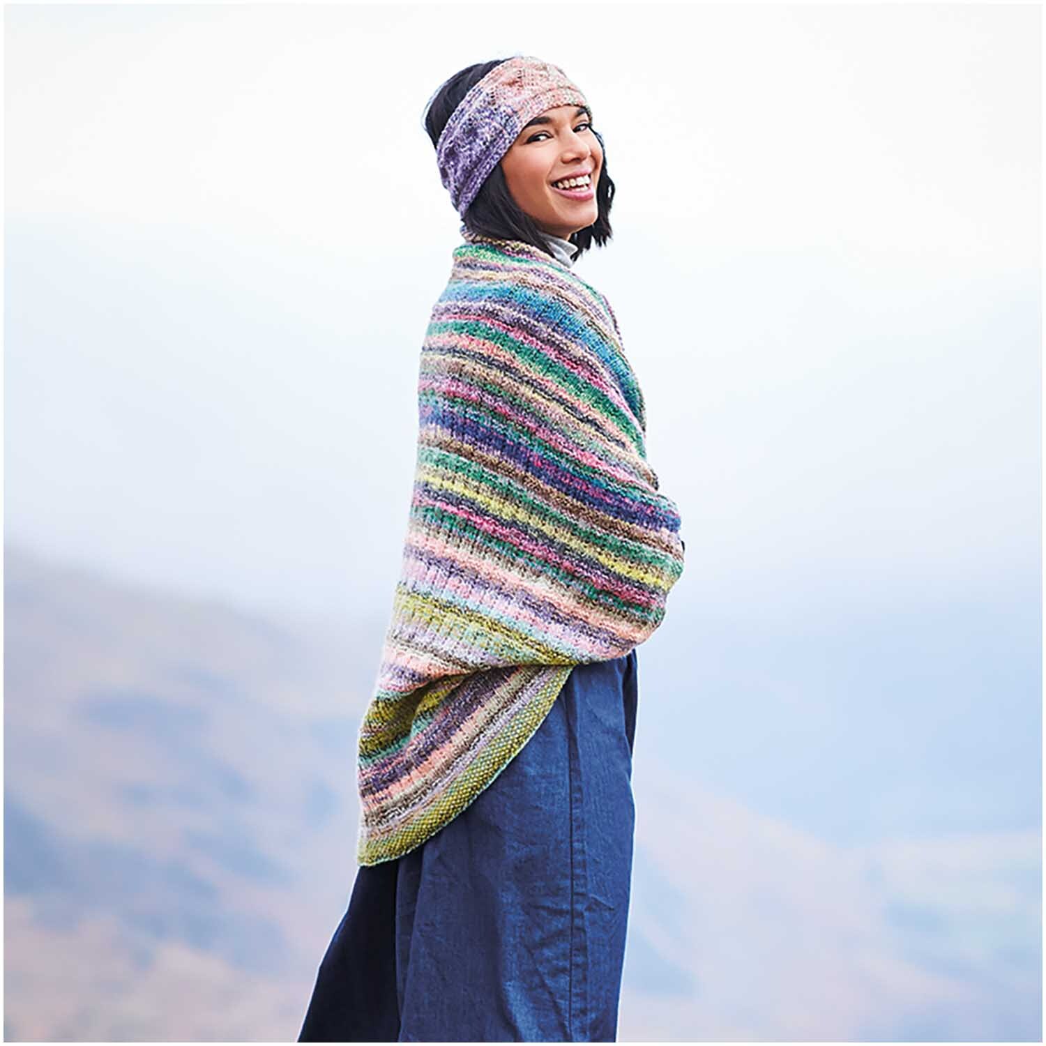 Strickset Poncho Modell 18: Frau trägt bunten Poncho und Stirnband vor nebliger Berglandschaft.