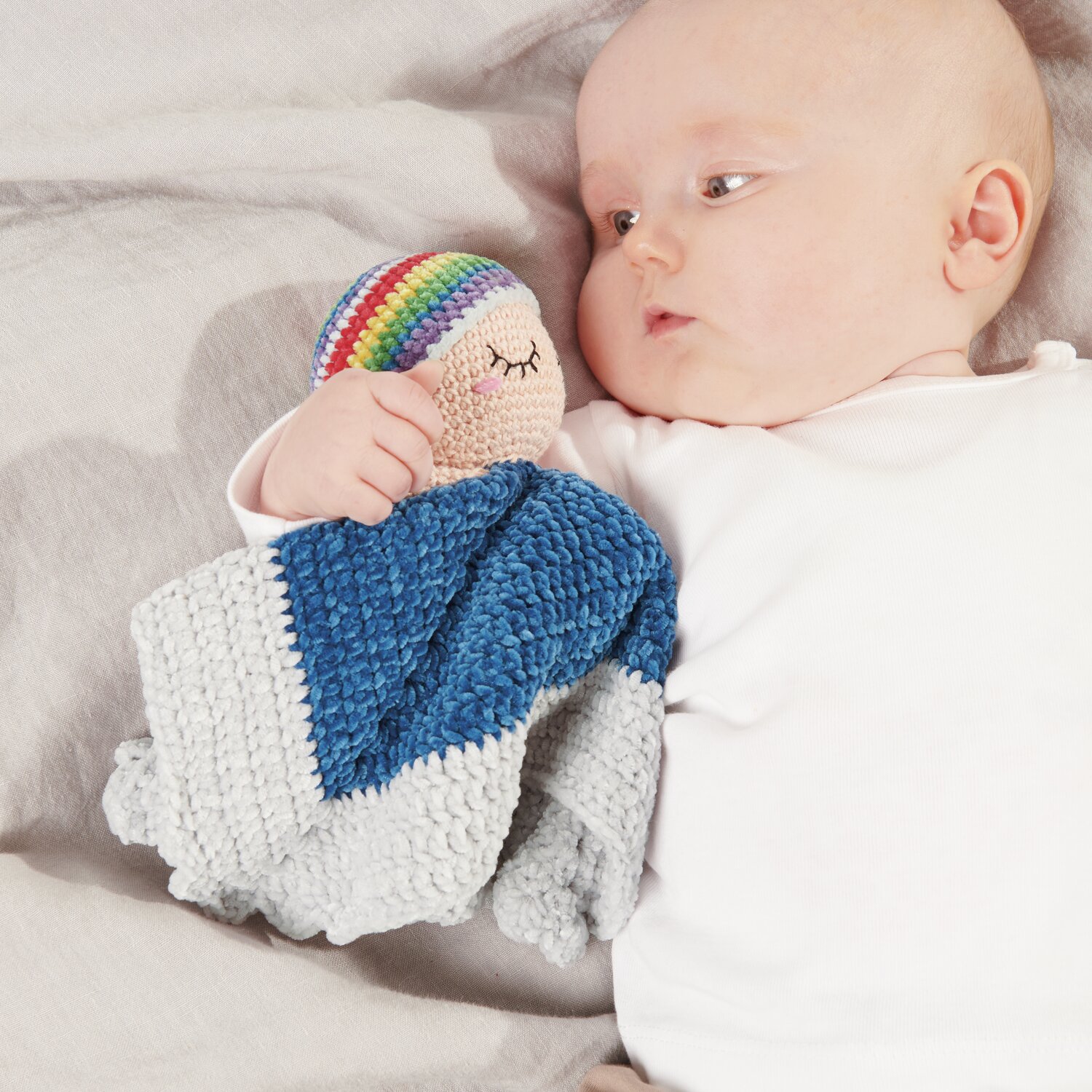 Ricorumi Baby Blankies Nilli Nilli: Ein Baby hält eine bunte Häkeldecke in Blau und Silbergrau von Rico Design.