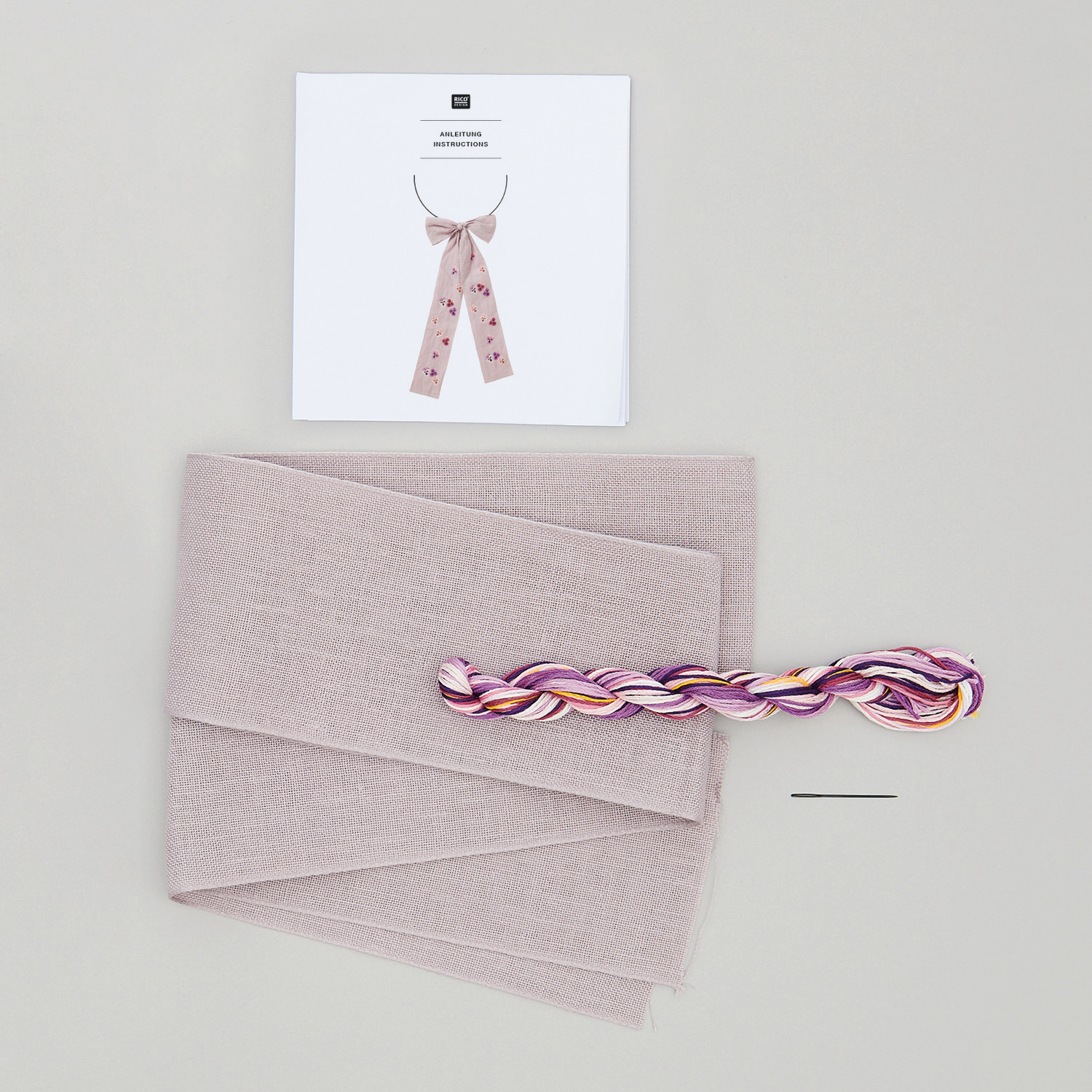 Stickpackung Schleife Stiefmütterchen von Rico Design mit rosa Stoff, Garn und Nadel auf grauem Hintergrund.