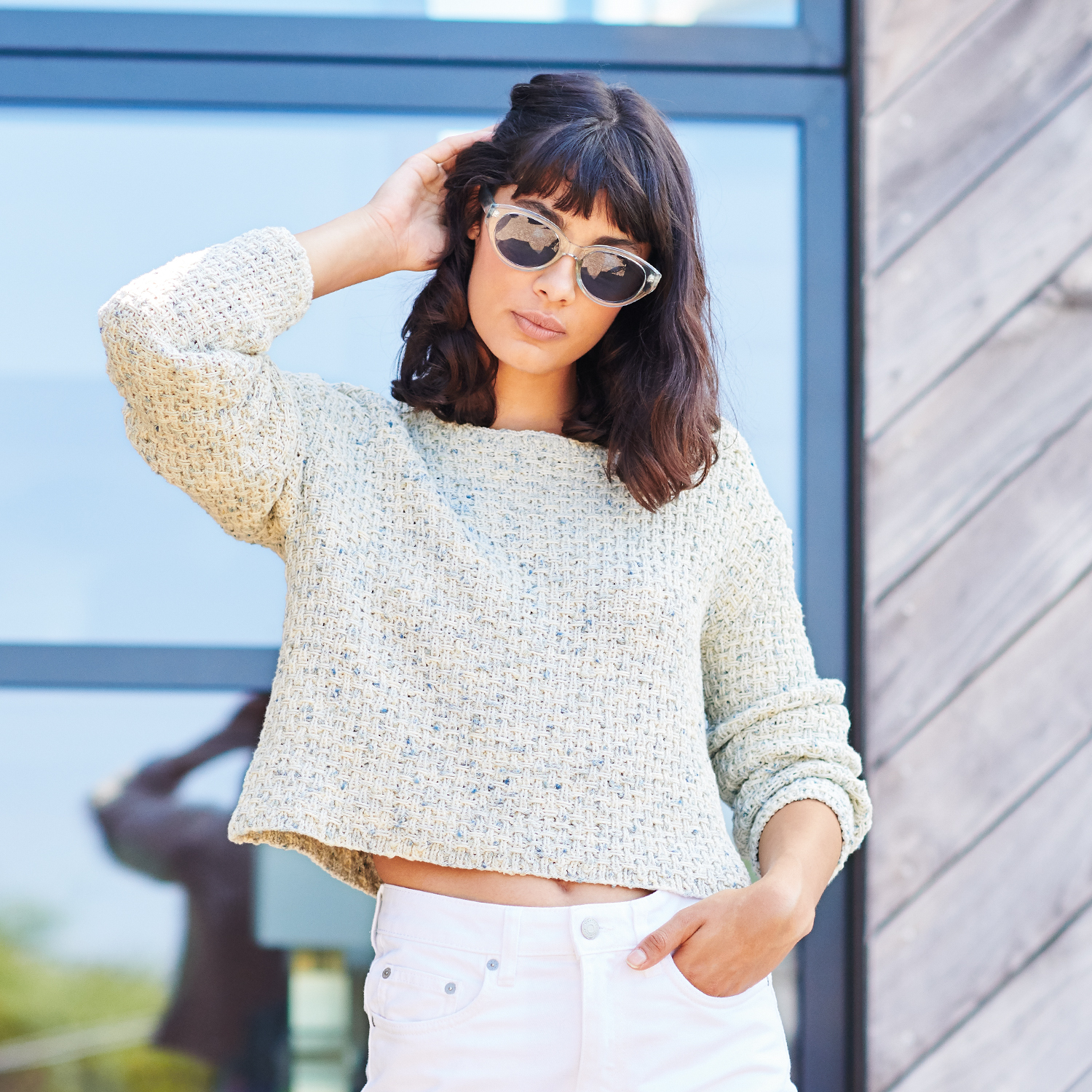 Strickanleitung Pullover aus Fashion Cotton Neps dk: Frau trägt hellen Strickpullover und Sonnenbrille vor modernem Gebäude.