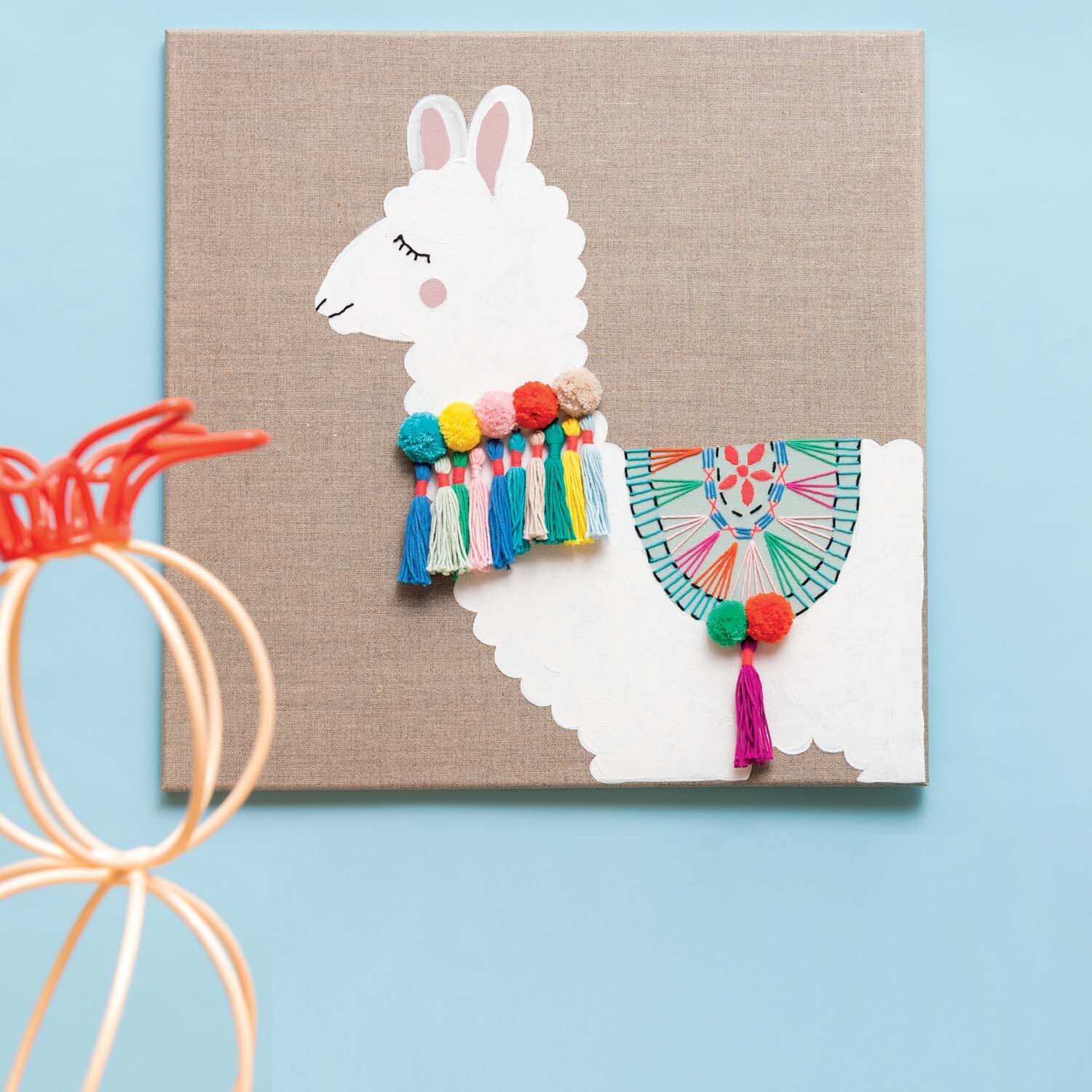Bastelanleitung Lama auf Leinwand: Ein weißes Lama mit buntem Pompon-Schmuck auf beigem Hintergrund.