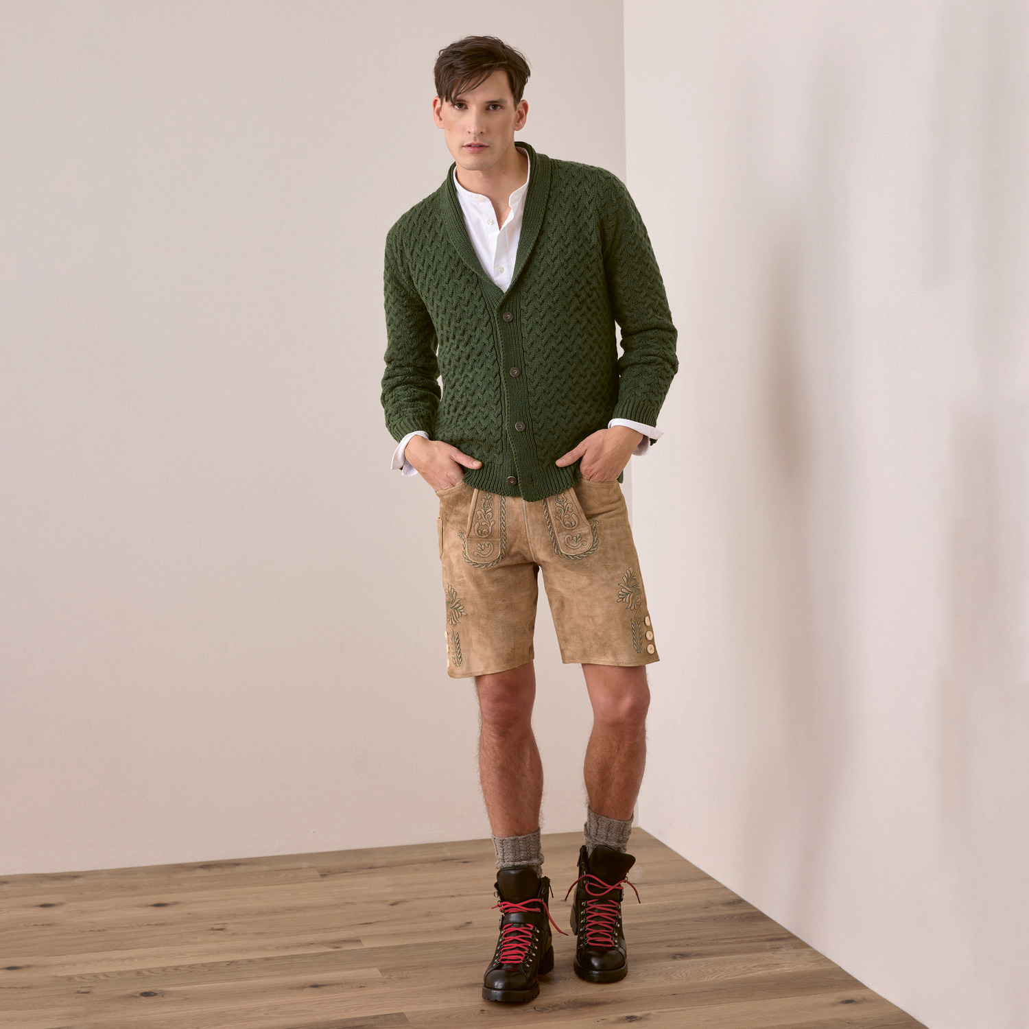 Strickset Herrenjacke Modell 27 aus Trachtenbuch I mog di: Mann in grüner Strickjacke, beiger Lederhose und Wanderschuhen vor weißer Wand.