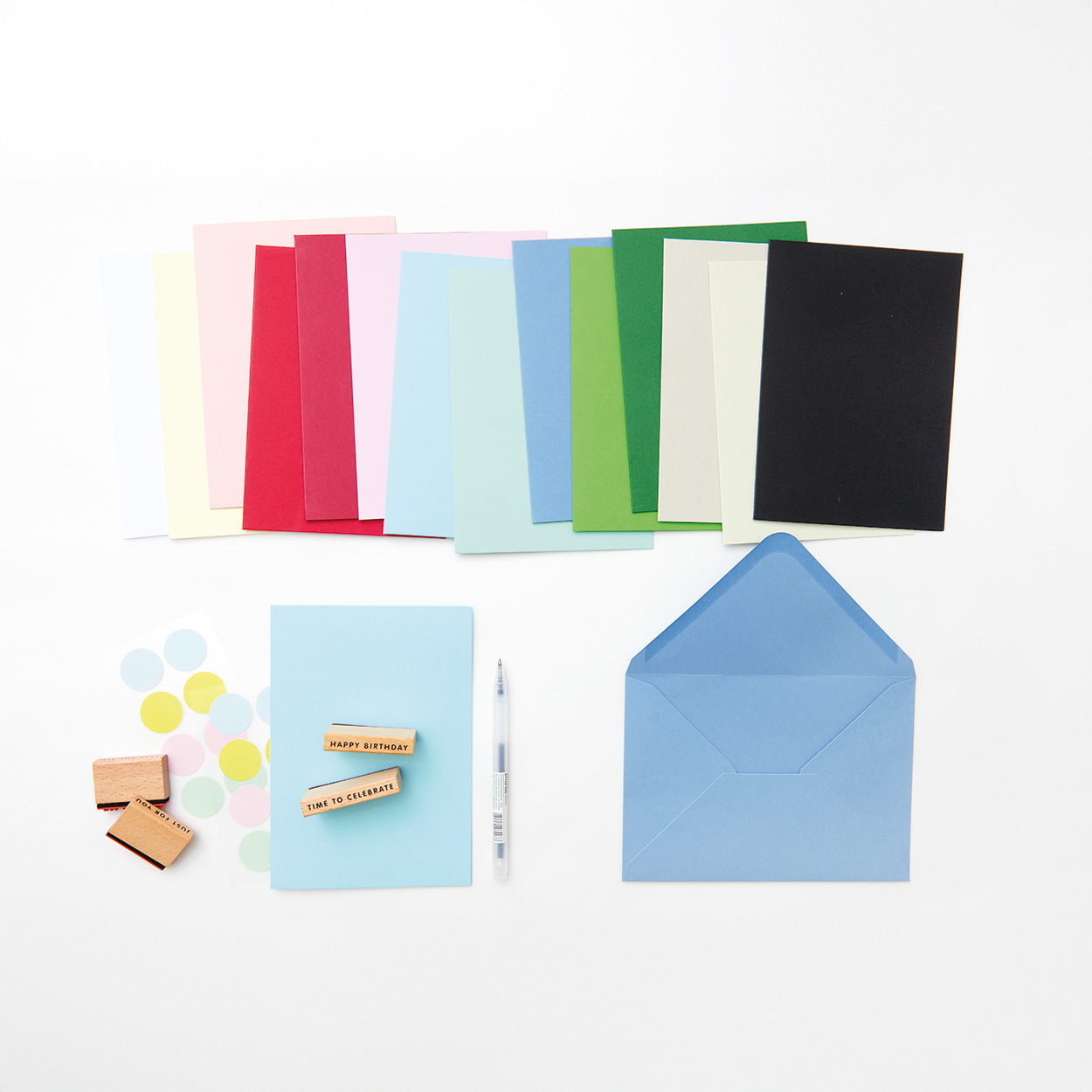 Basic Karten nach Farbe sortiert aufgereiht, darunter eine blaue Karte mit Stempel und Stickern, sowie ein blauer Umschlag. Flatlay von Produkten.