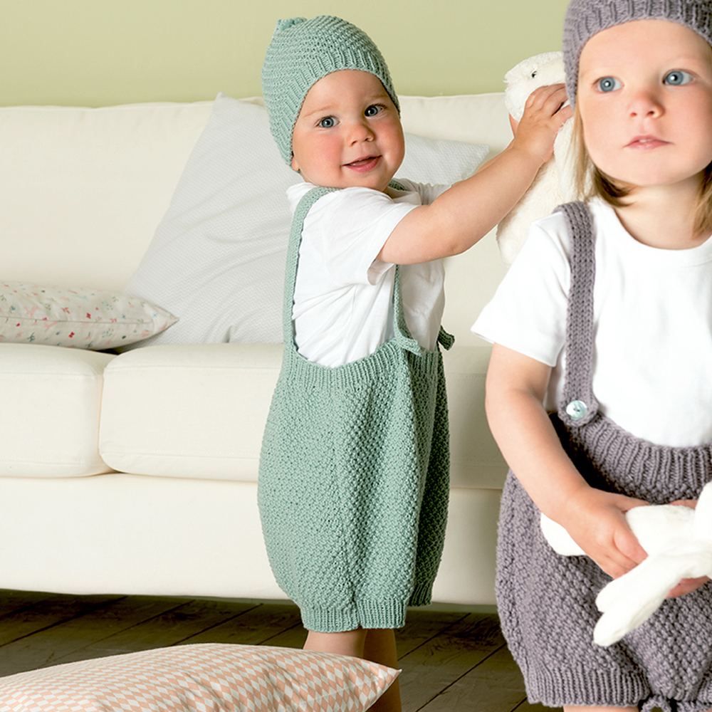Strickanleitung Baby Mütze, Hose & Booties: Zwei Kleinkinder in gestrickter Kleidung spielen auf einem Sofa.