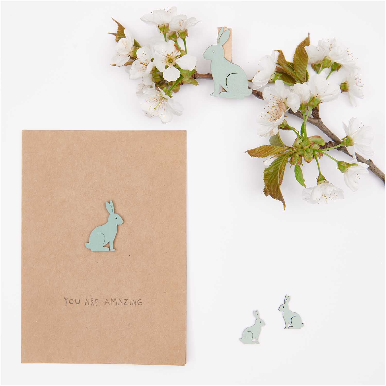 Dekoklammern Hase 6 Stück von Rico Design in Mint neben einer Karte und blühenden Zweigen.