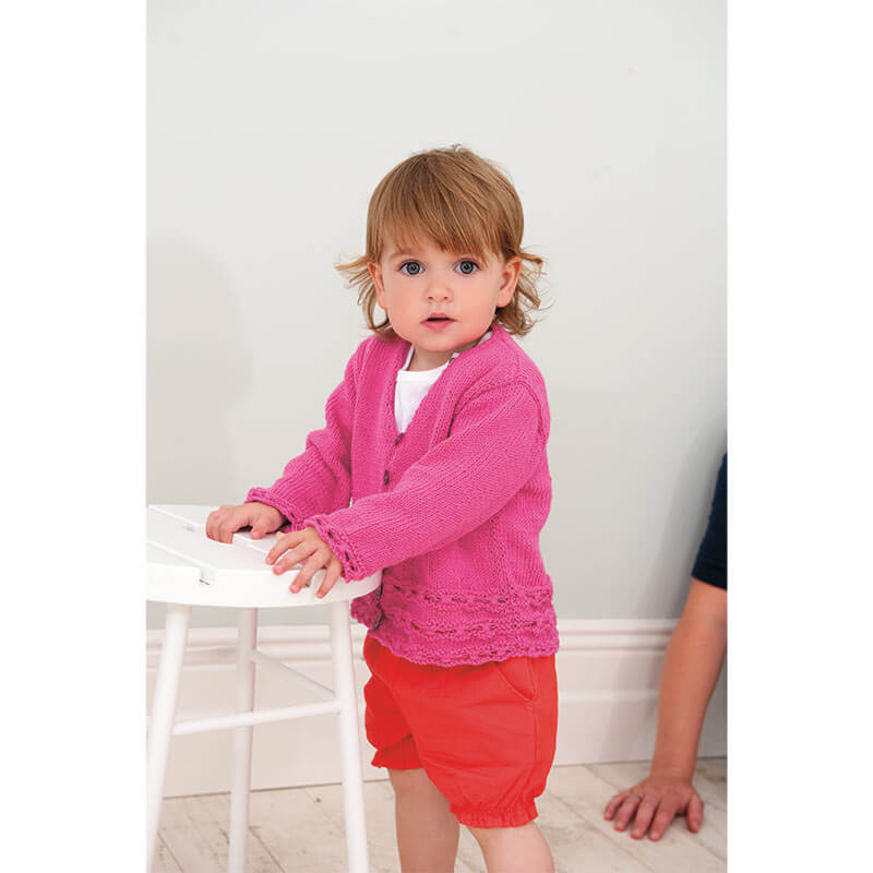 Rico Baby Heft Nr.9: Kleinkind in rosa Strickjacke und roten Shorts steht neben einem weißen Stuhl.