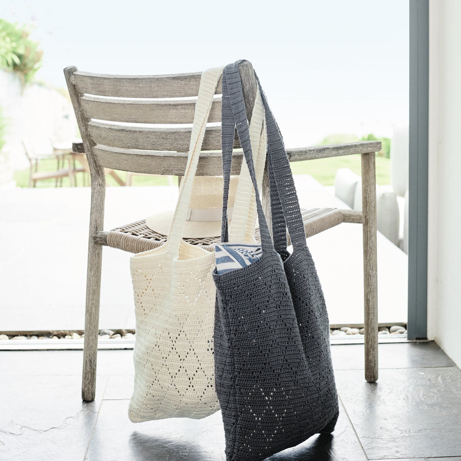 Häkelset Tasche Modell 06 aus Lovewool No. 20: Zwei gehäkelte Taschen in Creme und Grau hängen an einem Holzstuhl vor einem Fenster mit Gartenblick.
