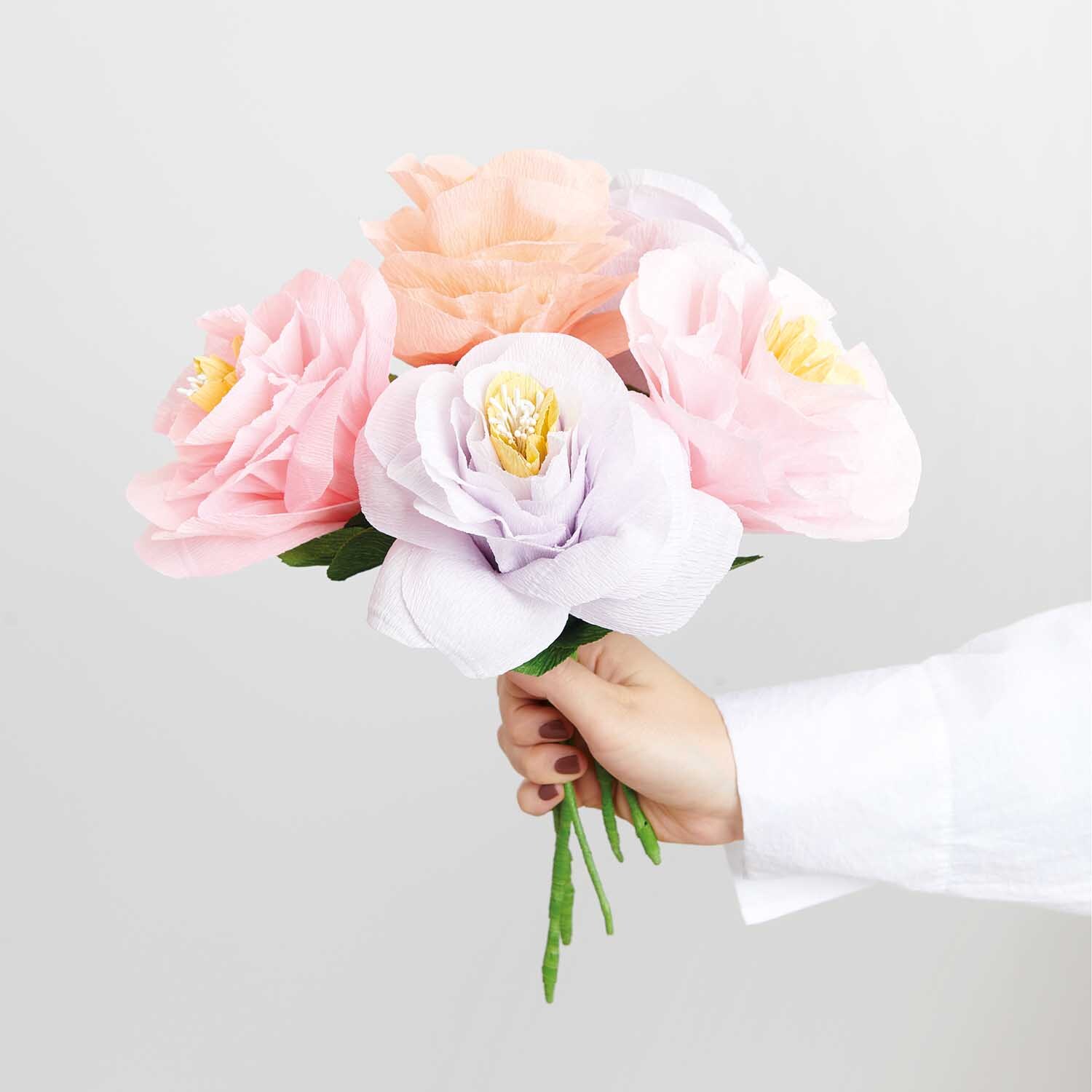 Bastelanleitung Papierblumen: Hand hält bunte Papierblumen in Pastelltönen vor weißem Hintergrund.