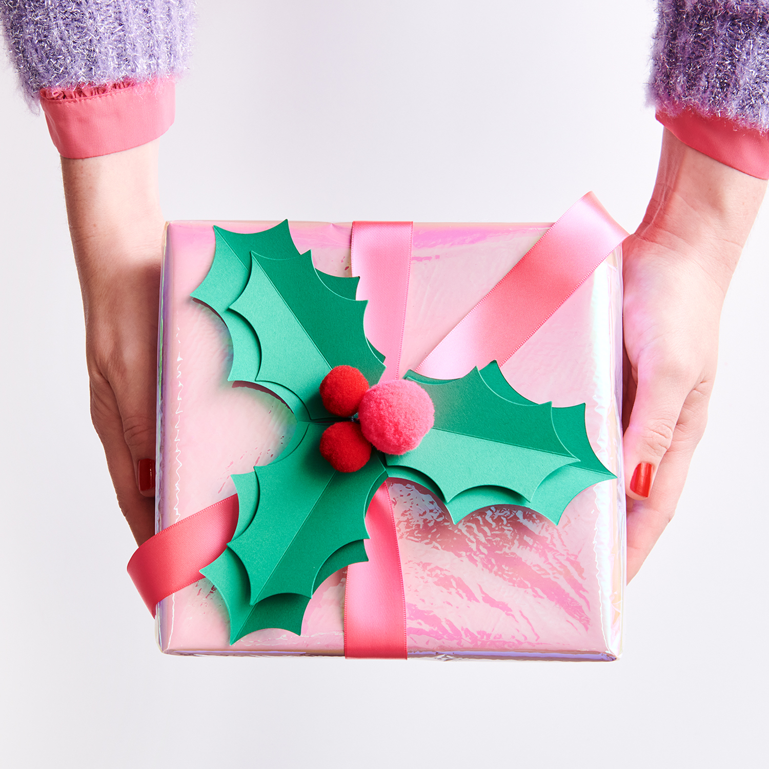 Bastelanleitung Ilex-Geschenkverzierung: Ein Geschenk mit rosa Schleife und grüner Ilex-Deko in Händen gehalten.