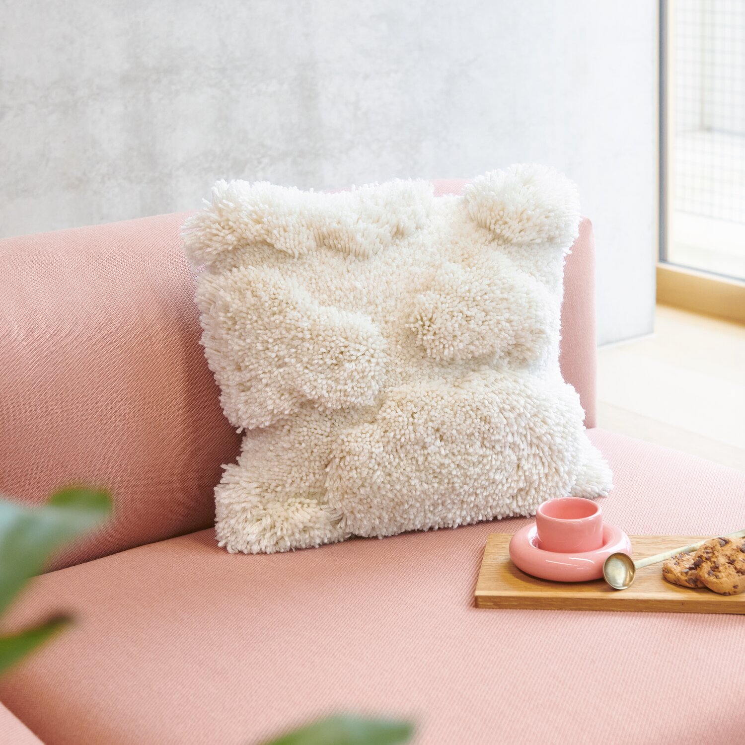 Rico Hooki Hooki Kissen aus flauschigem Knüpfgarn in Offwhite auf rosa Sofa, neben rosa Tasse und Keksen.