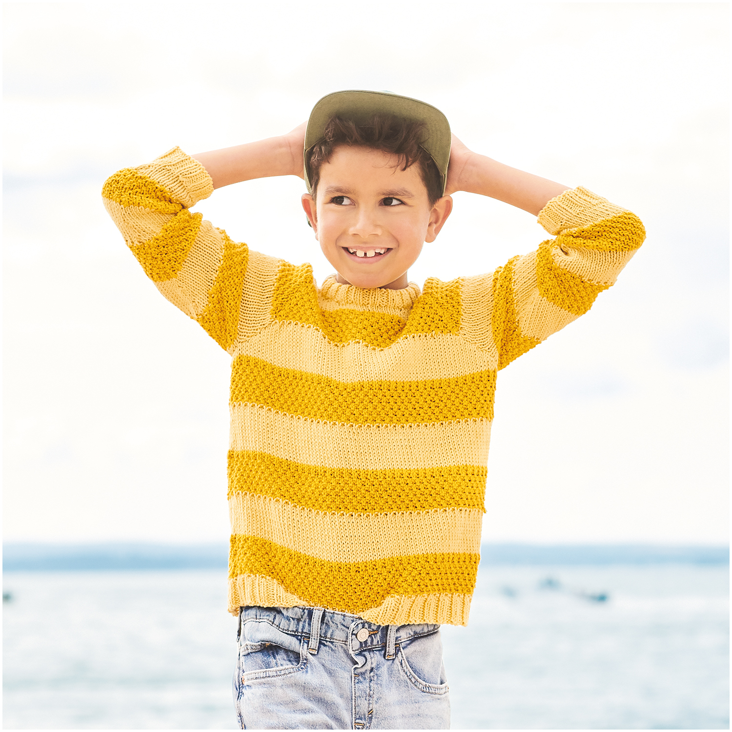 Essentials Organic Cotton aran: Junge in gestreiftem senfgelbem Pullover von Rico Design am Strand.