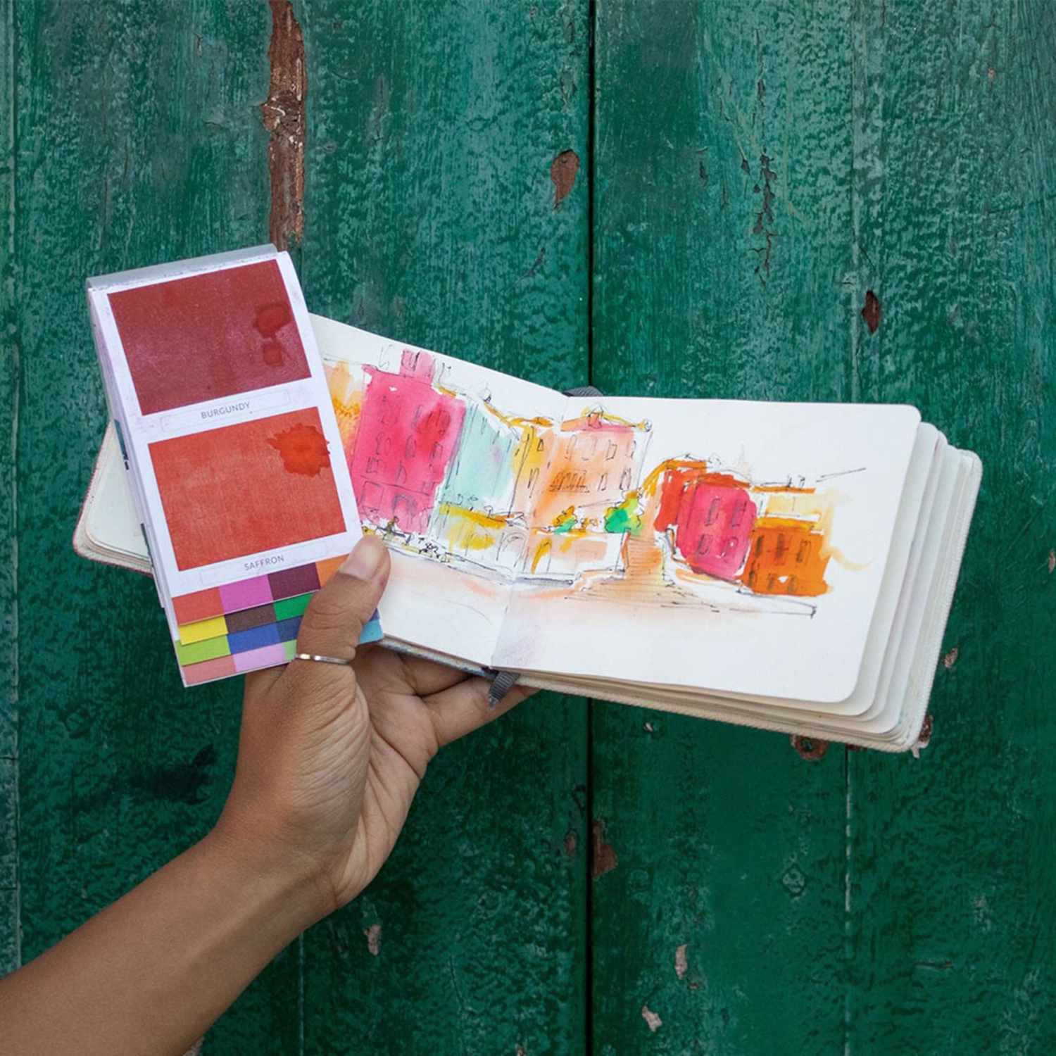 Viviva Colorsheets Spring: Hand hält Aquarellfarben und Skizzenbuch vor grünem Holzhintergrund. Ideal für kreative Künstler.