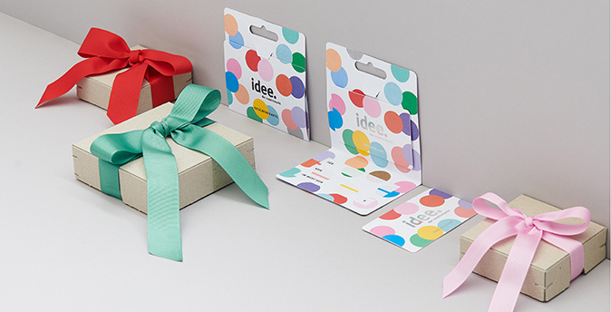 Zwei idee-Geschenkkarten an eine Wand gelehnt und eine liegend, sowie drei Geschenkboxen mit bunten Schleifen. Fotosetting graue Ecke.