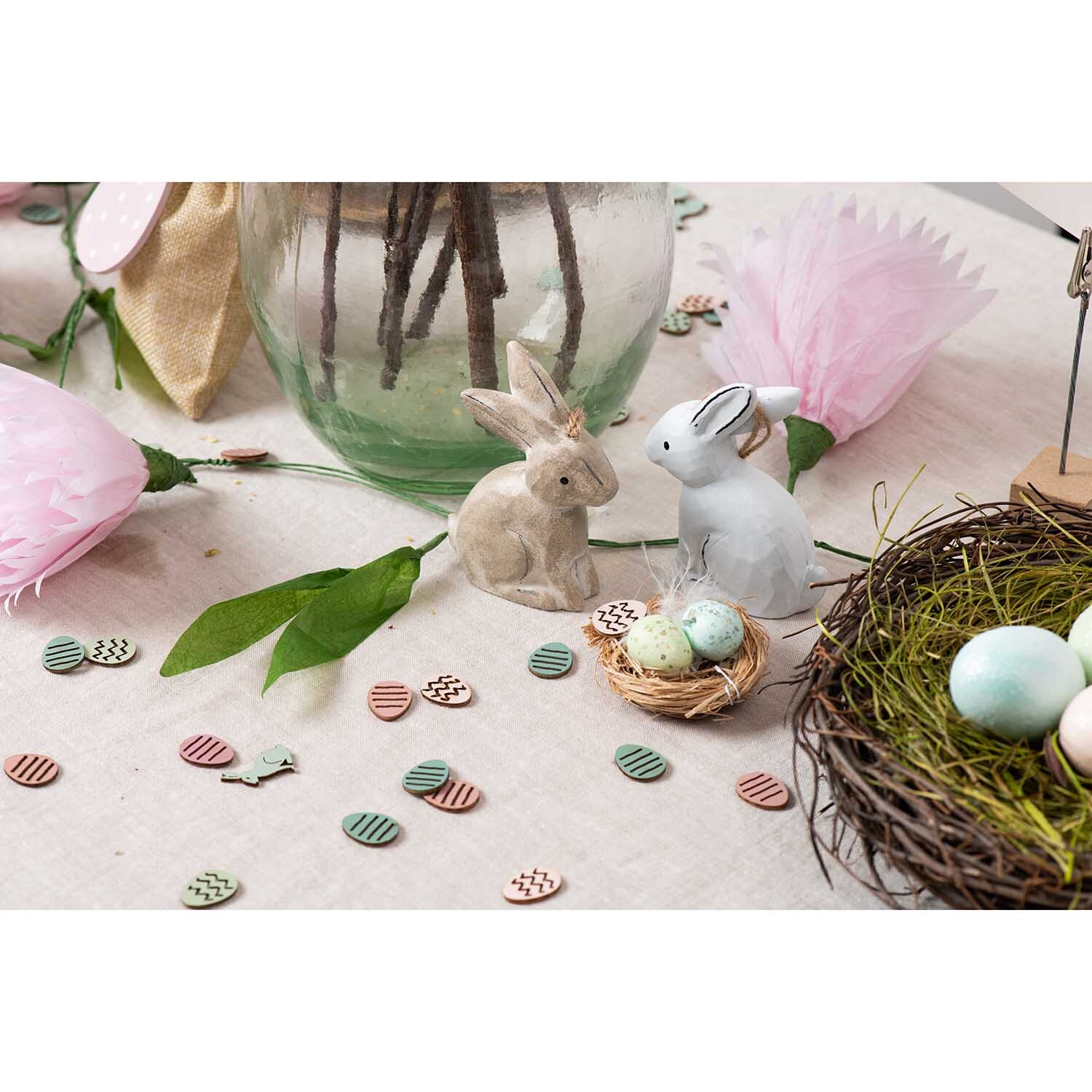 DIY Set Osterdeko pastell: Zwei Hasenfiguren neben einem Nest mit Eiern, umgeben von pastellfarbenen Dekorationen.