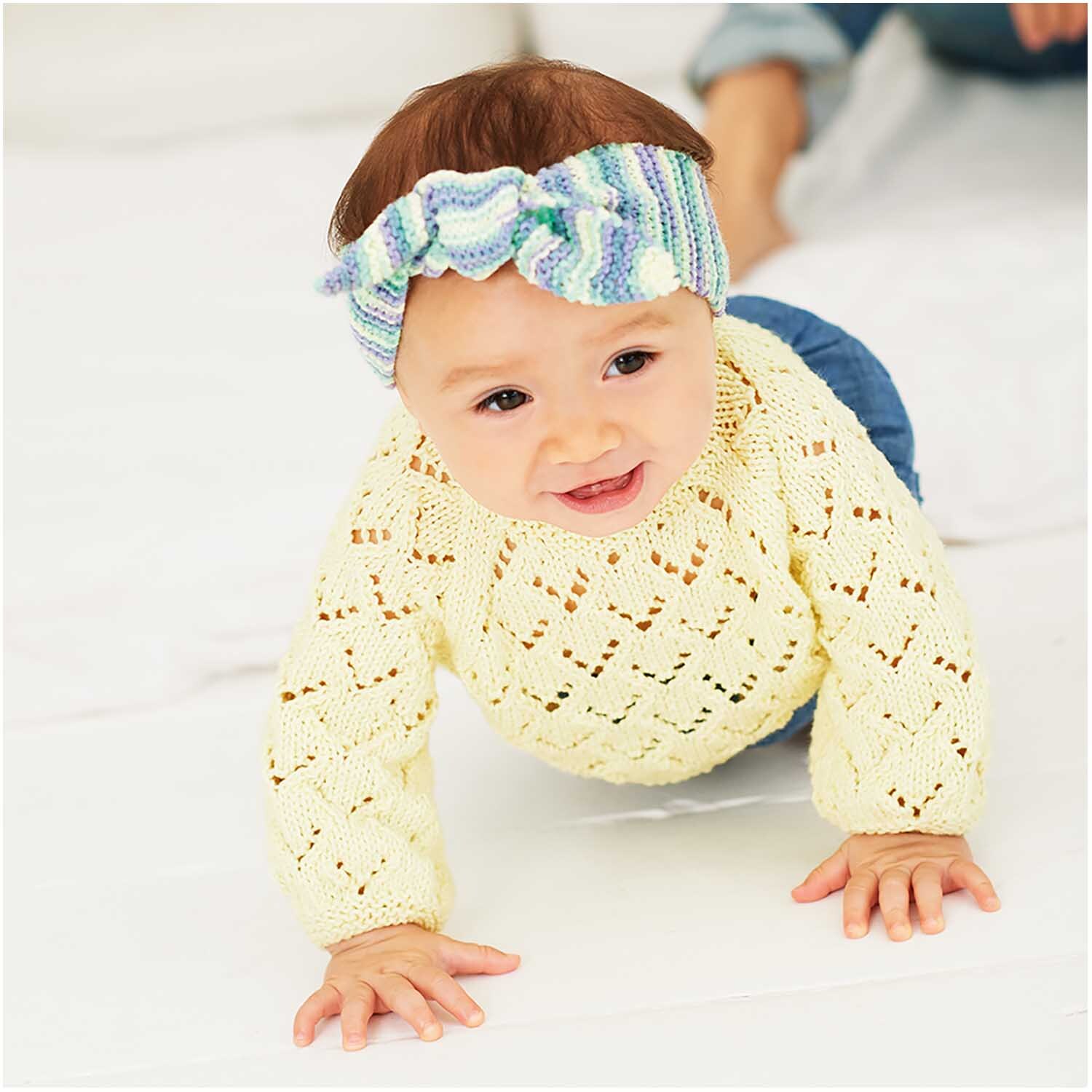 Rico Baby Cotton Soft Print dk: Ein Baby trägt einen gestrickten, gelben Pullover und ein Stirnband in Veilchen-Grün. Ideal für weiche Babybekleidung.