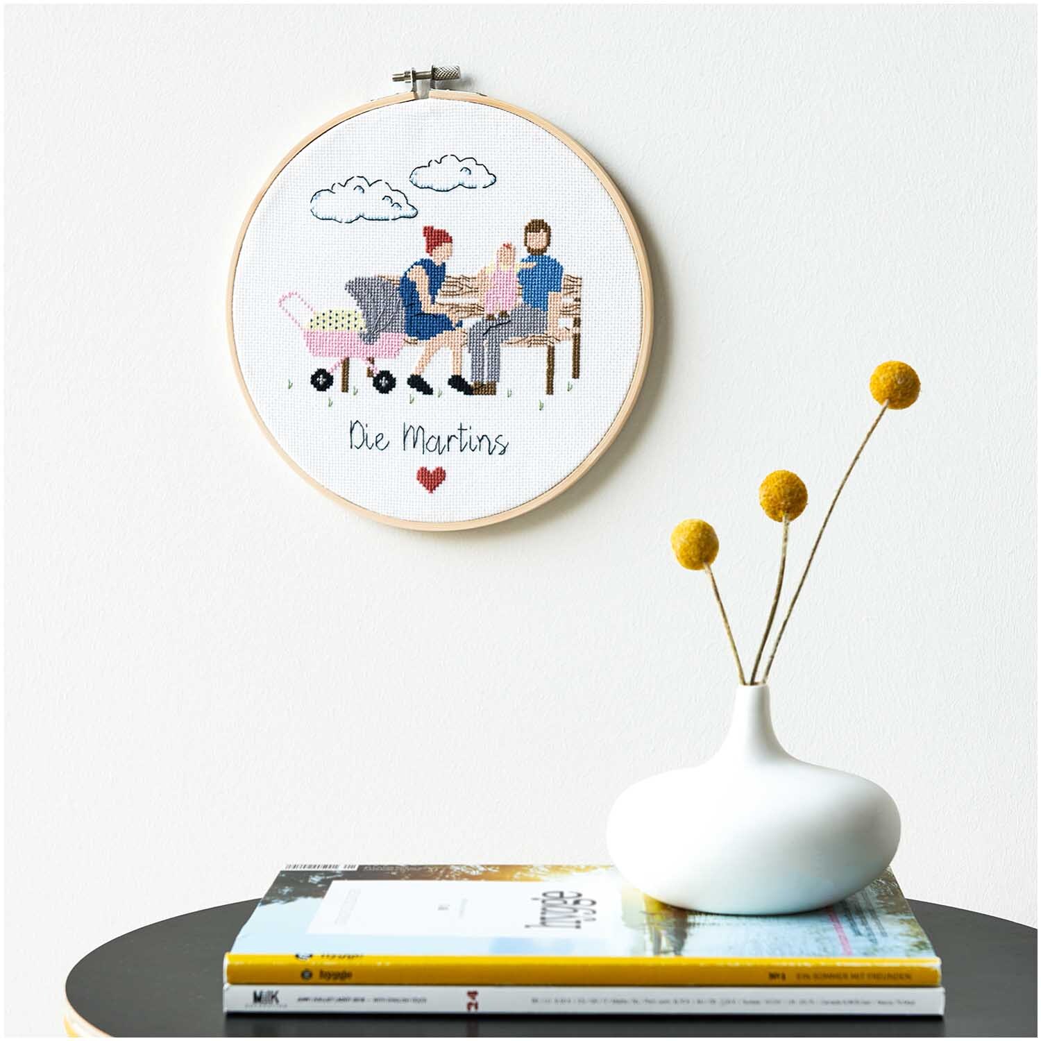 Figurico Stickpackung Young Family 20cm zeigt eine gestickte Familie auf weißem Stoff in einem Holzrahmen.