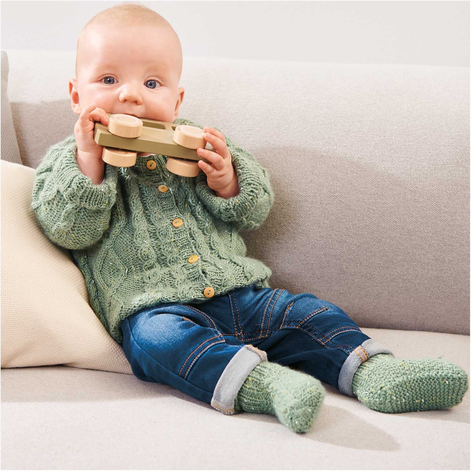 Rico Baby Dream Tweed dk - Ein Baby in einem gestrickten Outfit spielt mit einem Holzspielzeug auf einem Sofa.