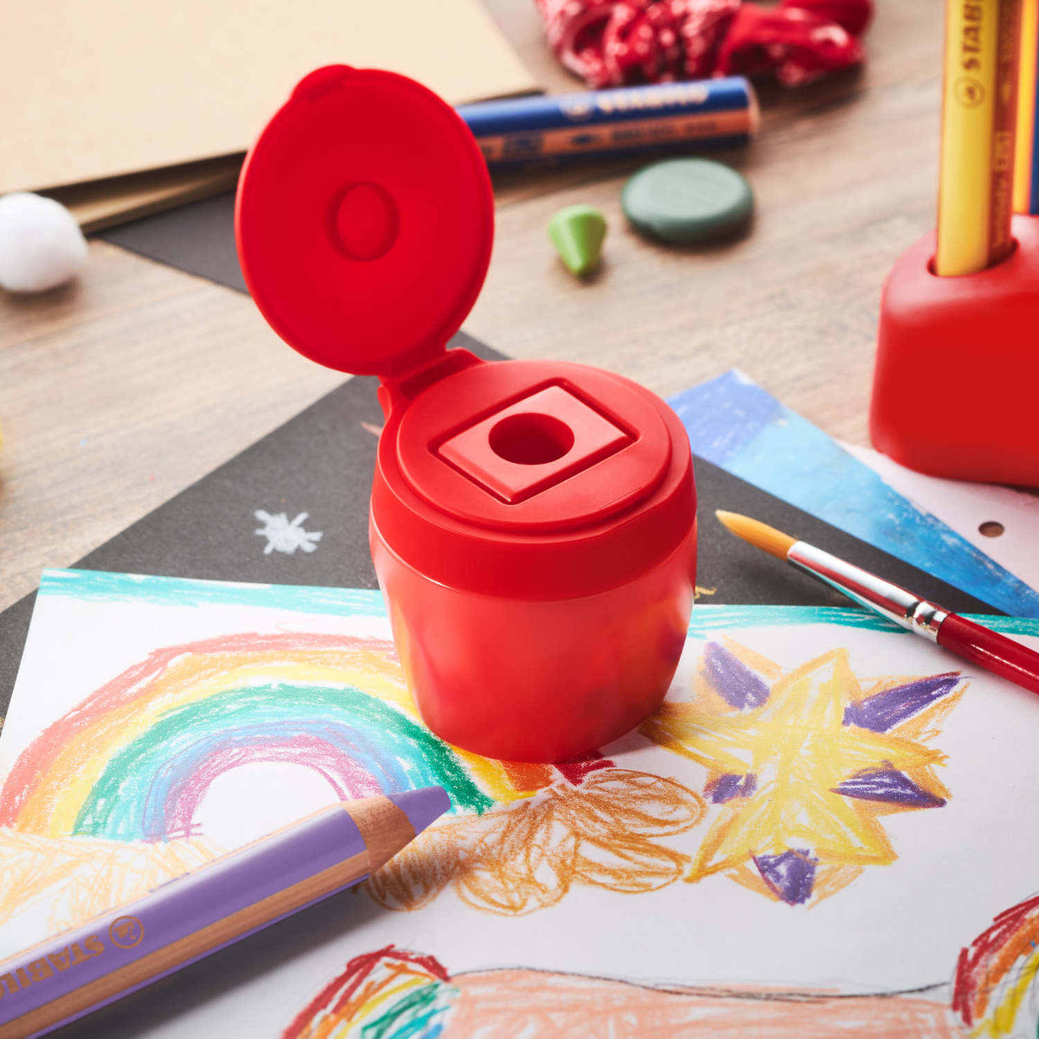 Stabilo woody 3 in 1: Roter Anspitzer auf einem Tisch mit Zeichnungen und Buntstiften im Hintergrund.