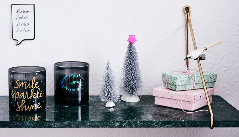 Anleitung Kerzengläser selber gestalten: Zwei dekorative Kerzengläser auf einem Regal mit Miniaturbäumen und Geschenkboxen.