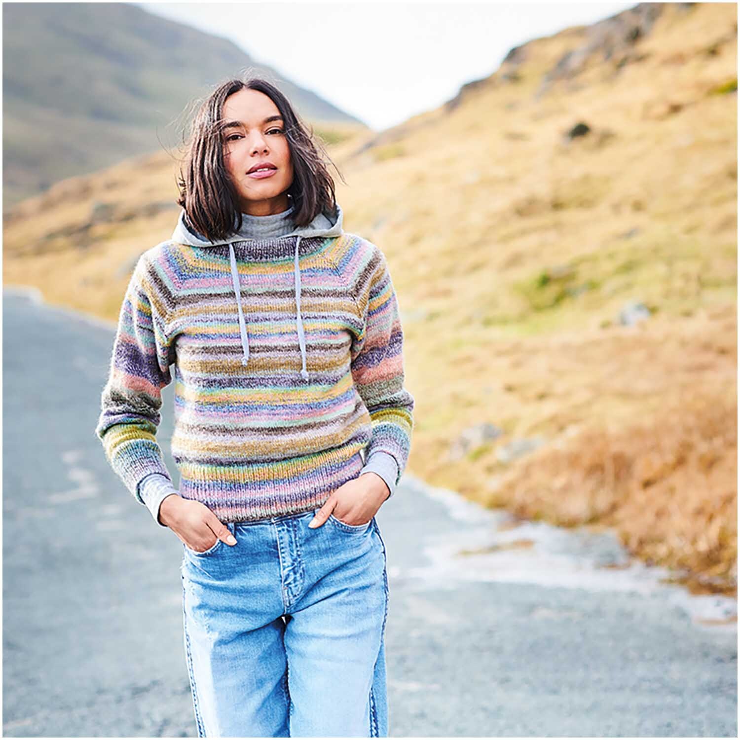 Strickset Pullover Modell 01: Frau in gestreiftem Pullover und Jeans auf einer ländlichen Straße.