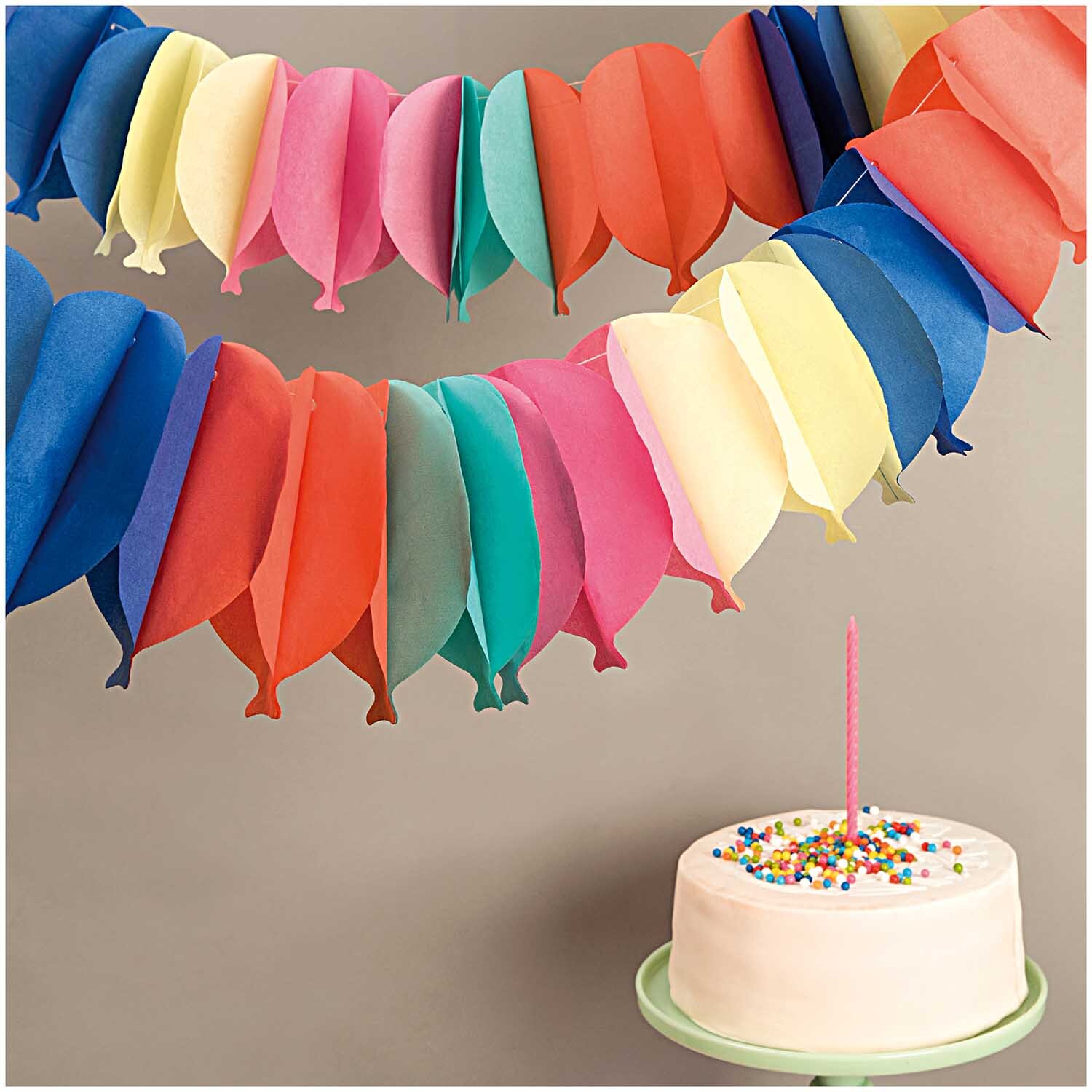 Wabenpapier Girlande Luftballons 6m in leuchtenden Farben von Rico Design, dekoriert über einem Kuchen mit Kerze.